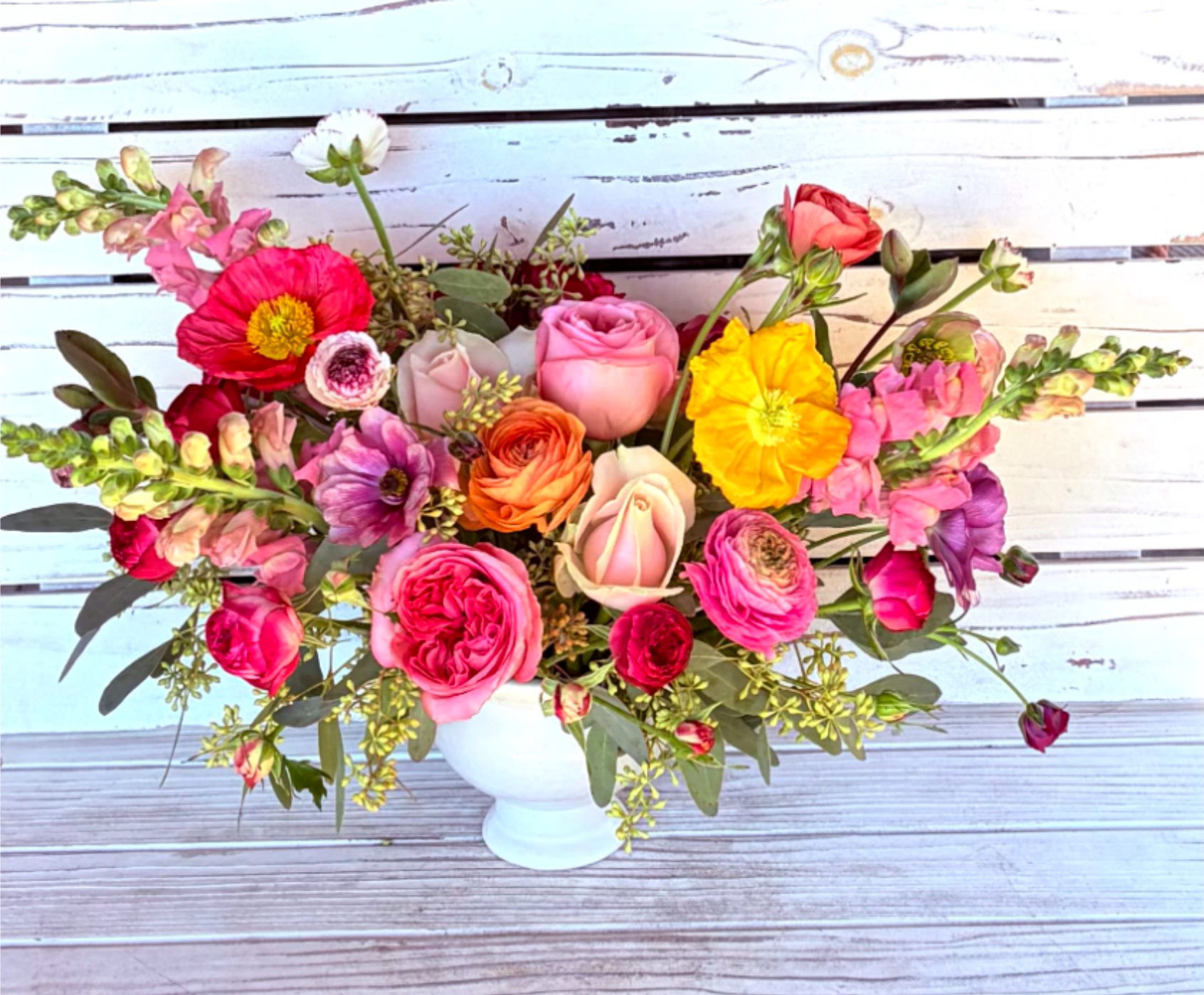 Colorful arrangement of roses, ranunculus, poppies, and snapdragons in a white pedestal vase