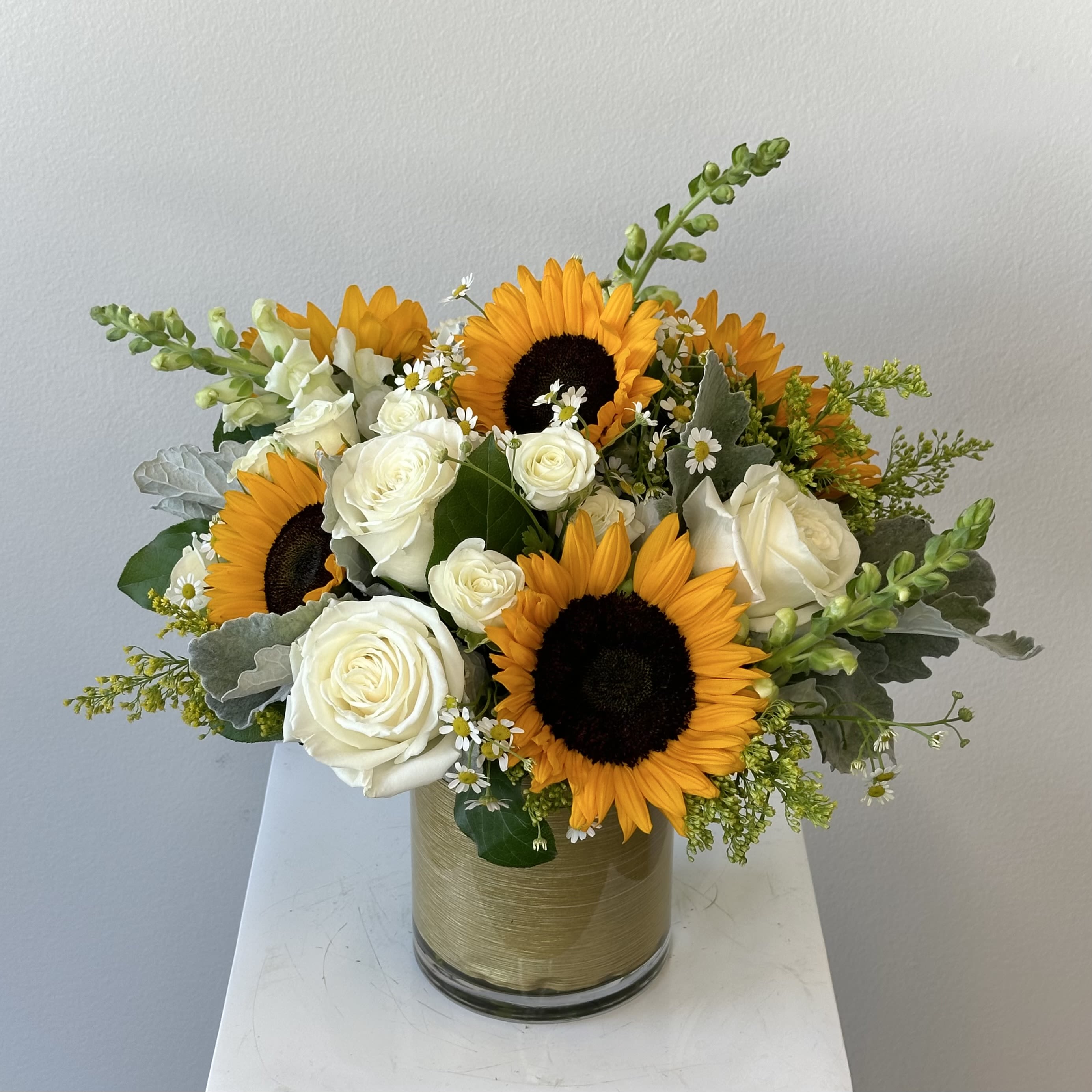 Golden Sunrise - Discover the radiant beauty of our Golden Sunrise flower arrangement, a stunning sunflower bouquet that captures the essence of a vibrant morning dawn. This luxurious floral masterpiece features bold, cheerful sunflowers as the focal point, symbolizing warmth, happiness, and positivity. Paired elegantly with delicate white roses for purity and romance, tall snapdragons adding height and texture in soft hues, and dainty chamomile blooms evoking calm and simplicity, this mixed flower bouquet creates a harmonious blend of colors and forms that's both uplifting and sophisticated. Presented in an exquisite gold-lined vase, the Golden Sunrise elevates any space—perfect for adding a golden glow to your home decor, tabletop centerpiece, or as a thoughtful gift. The sunflowers' large, sunny faces contrast beautifully with the creamy white roses and whimsical chamomile, while snapdragons provide vertical interest and a pop of subtle color, making this arrangement ideal for summer vibes, birthdays, anniversaries, thank you gestures, or simply to brighten someone's day. Handcrafted with fresh, high-quality blooms, our sunflower arrangement with white roses, snapdragons, and chamomile is designed to last. Whether you're searching for unique sunflower bouquets, mixed floral arrangements, or luxury flower delivery options, the Golden Sunrise is a timeless choice that radiates optimism and style.