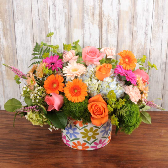 Low arrangement of orange and pink roses and gerbera daisies with blue hydrangea in a colorful round container