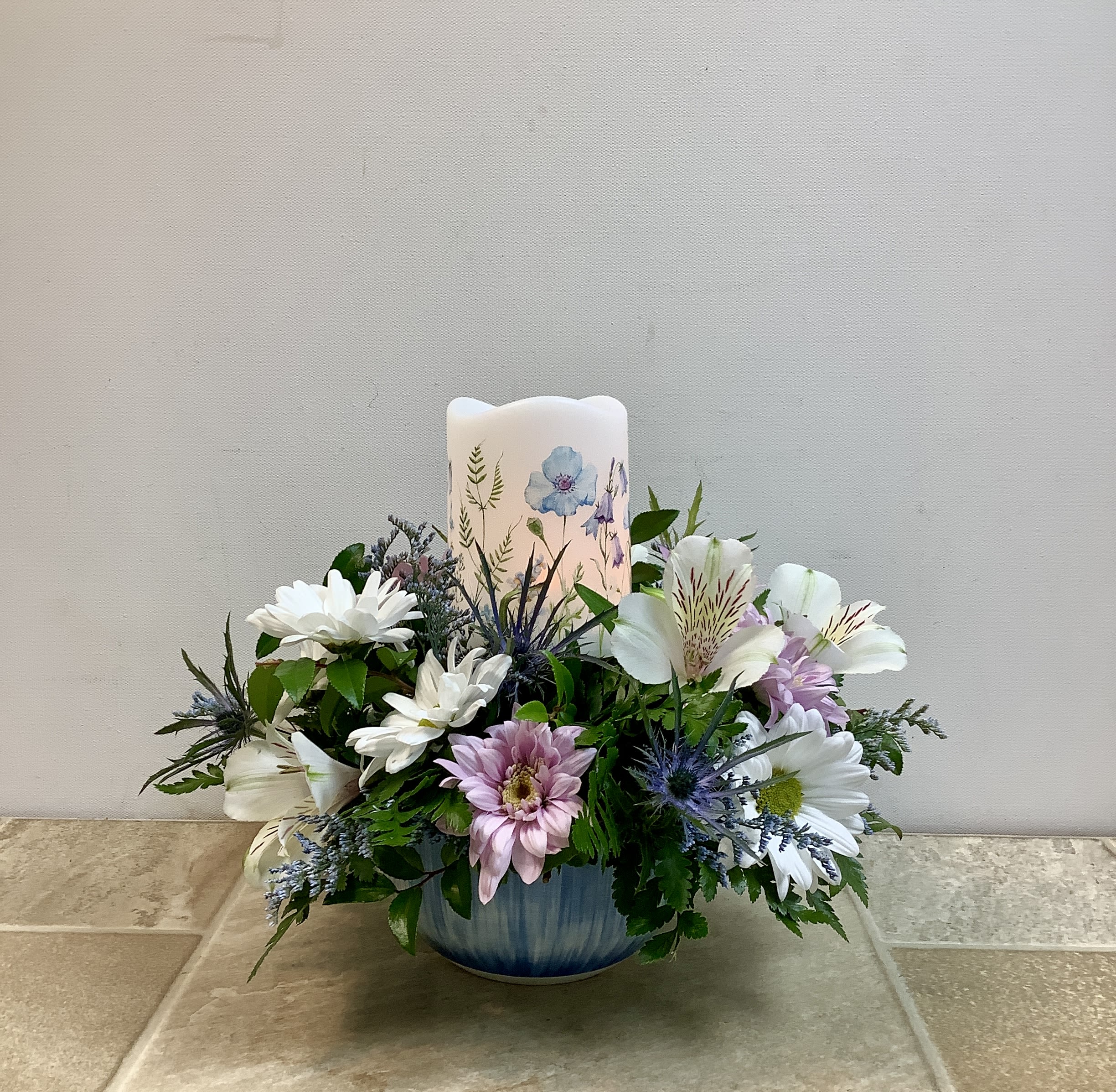 Candlelight - Soft, serenity-first styling sets this flower arrangement apart - a low, rounded candlelit centerpiece perfect as a sympathy arrangement, a quiet celebration gift, or a thoughtful everyday bouquet. The led pillar candle at the center casts a gentle glow that makes the white and lavender daisies, blue thistle and white alstroemeria feel even more tender, while cool blue accents in the ceramic bowl vase lend a peaceful, calming note. It measures 11” wide x 10” tall.  
