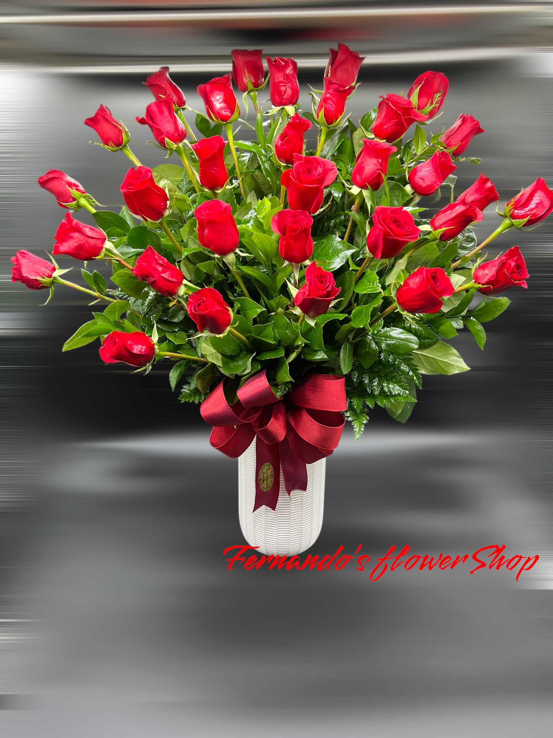 Tall white vase filled with many red roses and foliage, finished with a large red satin bow