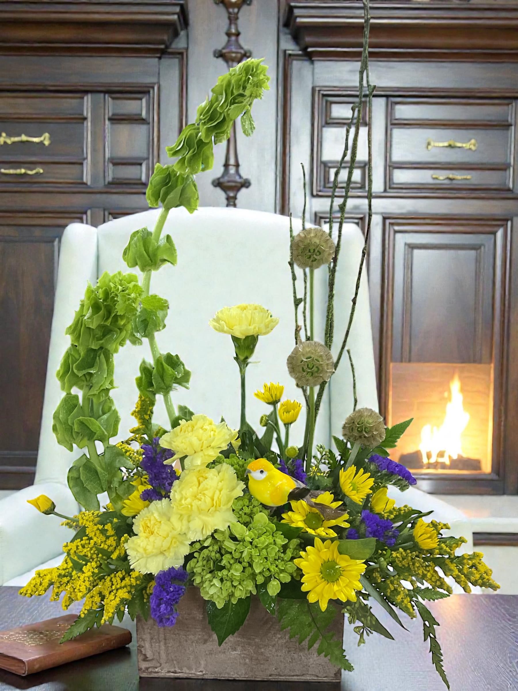 Sunlit Grace -  Sunlit Grace is a bright, garden-style arrangement designed with height, movement, and clean structure. Golden yellow roses and daisy poms are layered with airy purple statice and fresh greenery, creating a lively, balanced design that feels open and naturally uplifting.  Vertical lines of Bells of Ireland and curly willow draw the eye upward, giving the piece a light, sculptural shape, while textured greenery adds depth without crowding the design. Nestled naturally within the blooms, a small bird accent brings a soft, seasonal touch—subtle, intentional, and perfectly placed.  Thoughtfully handcrafted by Aspen Tower Florist, Sunlit Grace is a beautiful choice for birthdays, encouragement, thinking of you, or simply bringing a little brightness into the room.  Each arrangement is created with the freshest stems available. Varieties may vary slightly based on availability while maintaining the same color palette and overall design.  Available for local flower delivery through Aspen Tower Florist, serving Greensburg and Shelbyville along with Rushville, Batesville, Westport, Oldenburg, St. Paul, Waldron, Milroy, and Hope.