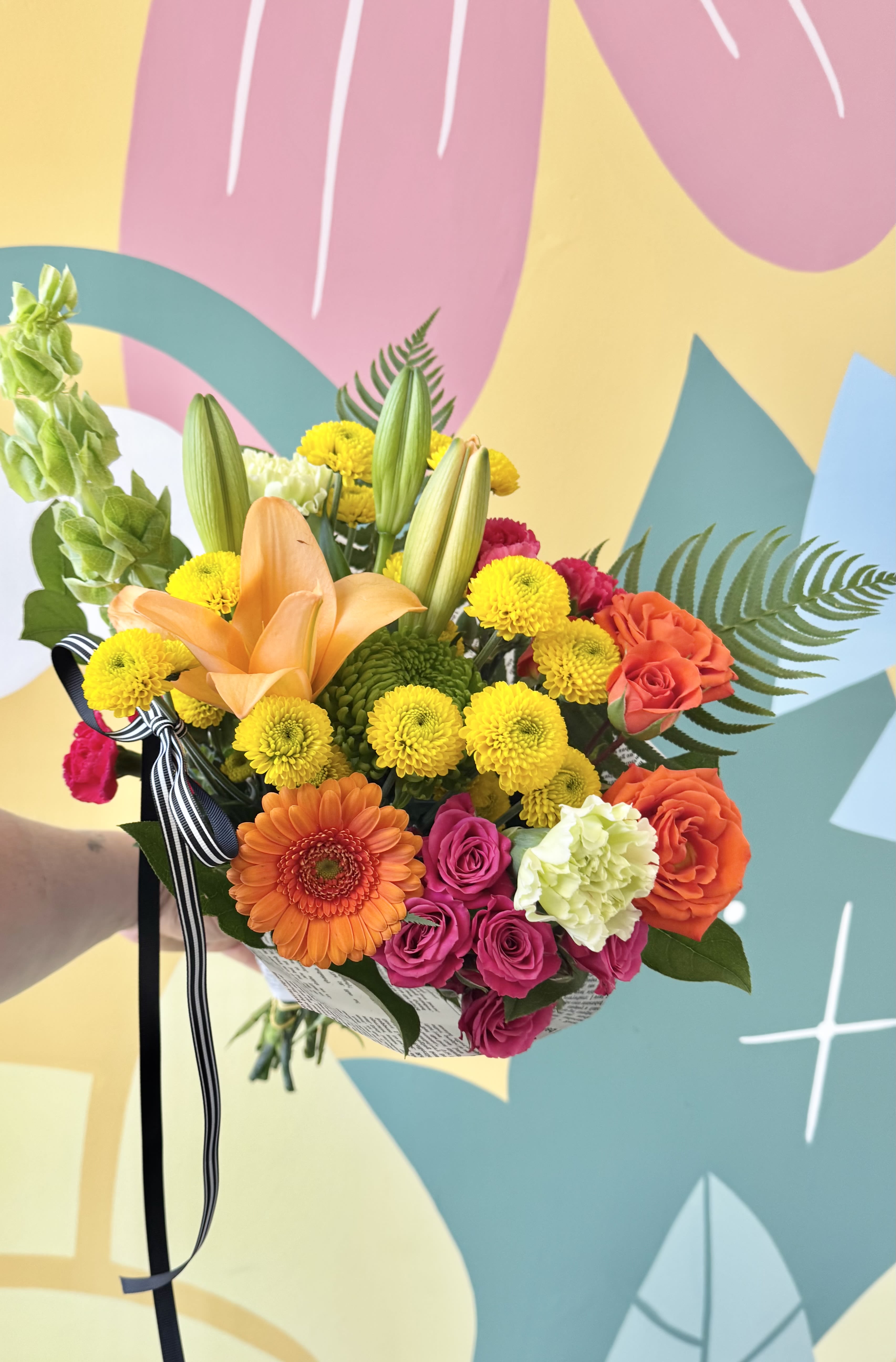 Bright and cheery hand tied bouquet  - Sunlit yellow chrysanthemums and vibrant orange lilies set a joyful tone in this bright and cheery hand tied bouquet, perfect for birthdays, congratulations, or any moment you want to celebrate. Coral and pink roses, a single orange gerbera daisy, pale green carnations and snapdragon accents build a lively, rounded arrangement that reads energetic and uplifting-the kind of bouquet that makes the person you're thinking of feel noticed and celebrated.  Full and hand-tied with several unopened lily buds for height and a fresh, just-picked look, this bouquet is finished with a black-and-white striped ribbon and a printed bouquet wrap for a polished, gift-ready presentation. The mix of warm yellows, pinks, oranges and soft white highlights creates strong visual contrast and a sunny, textured feel that works equally well on a table centerpiece or as a doorstep surprise.