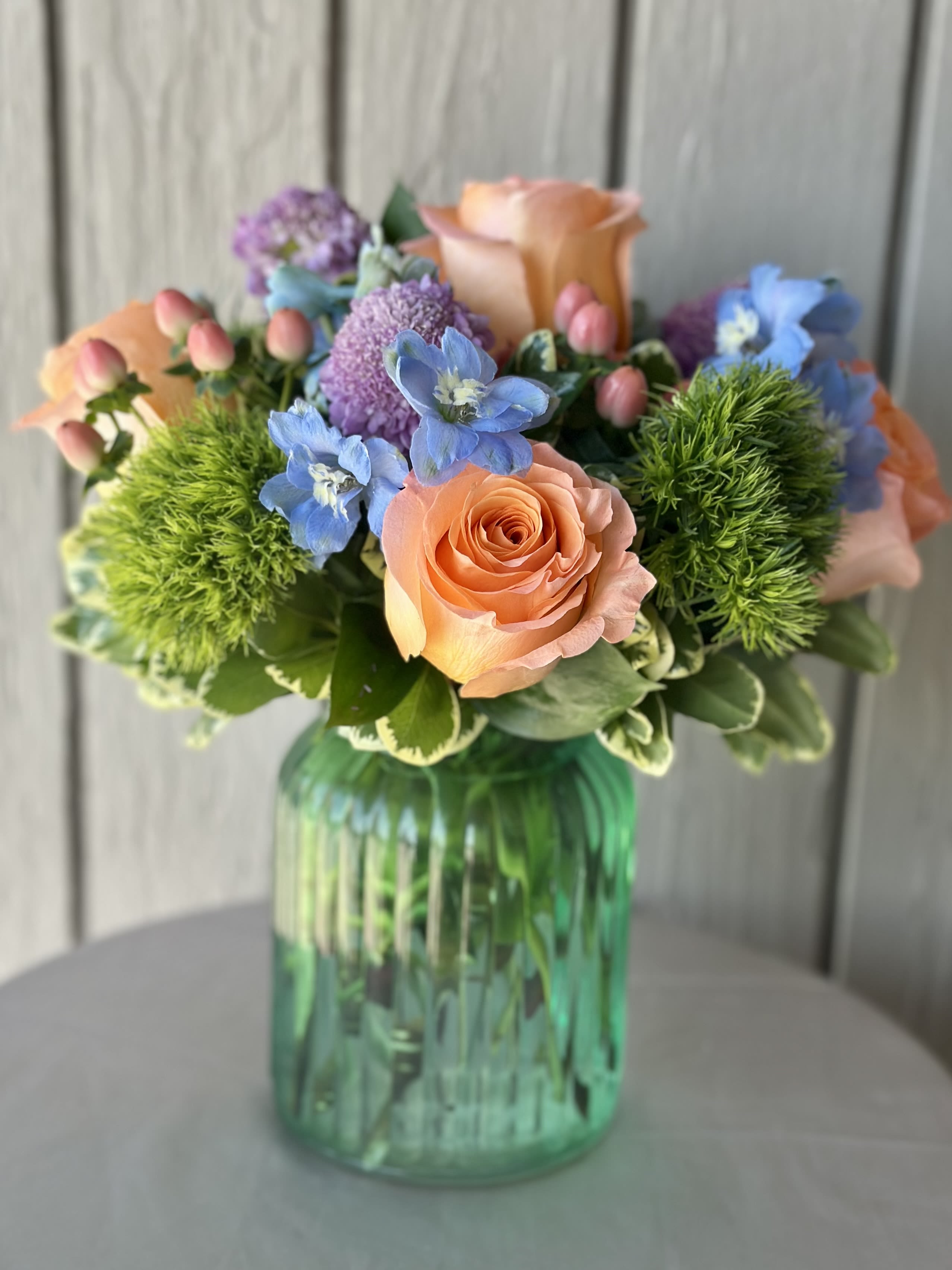 Meadow Fresh  - Peach roses, blue blooms and pink buds in a green glass vase-bright, playful mixed-color bouquet perfect for birthdays, thank-yous, or cheerful celebrations.