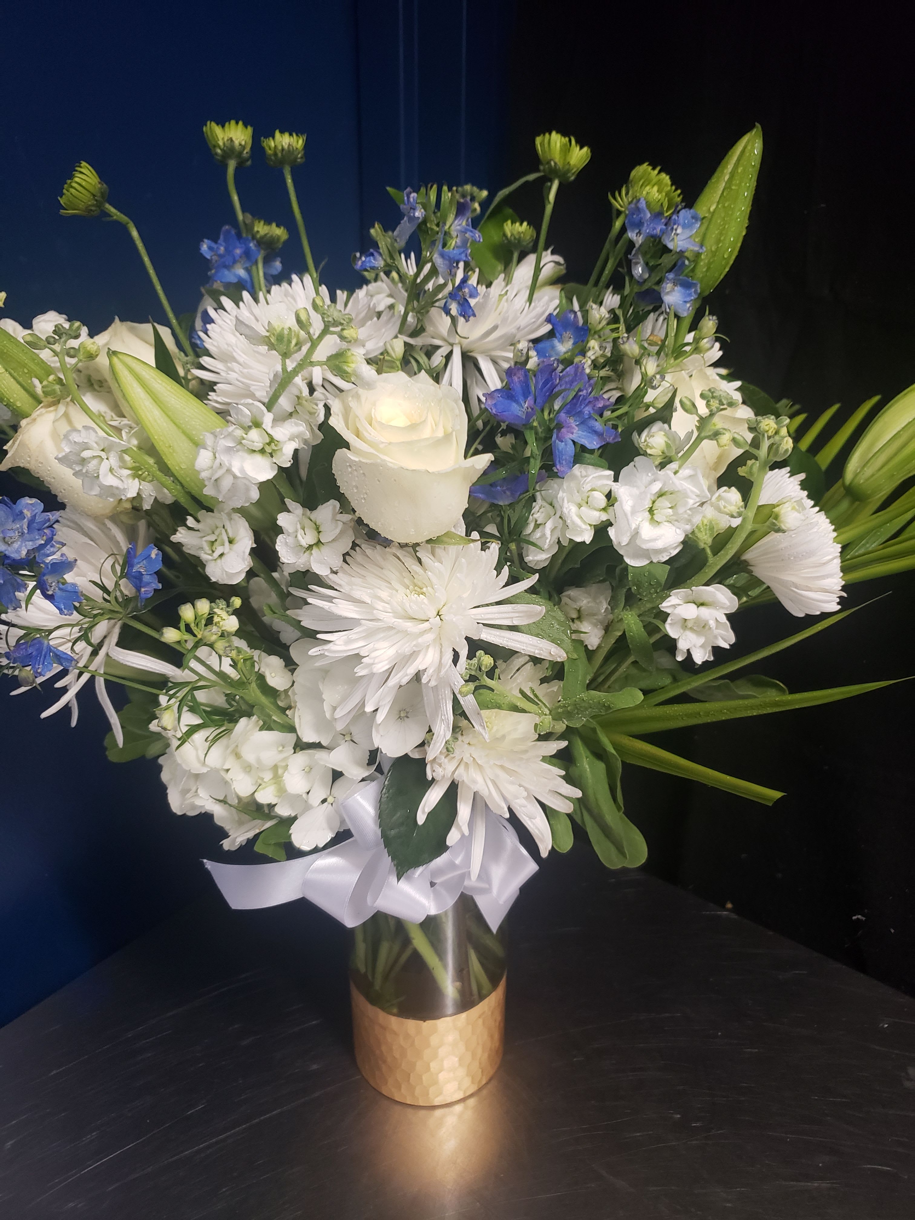 LOVE WHITE AND BLUE - Pure white blooms framed with bright blue accents create a calm, uplifting statement-perfect as a sympathy arrangement or a refined wedding centerpiece. The bouquet's white lilies, roses, and chrysanthemums form a full, rounded silhouette while vivid blue delphinium-like flowers and fresh green buds add airy texture and a cool-toned contrast that feels both respectful and celebratory. It reads as elegant and serene, the kind of gift that makes the person you're thinking of feel comforted and seen.  Arranged in a clear glass vase and finished with a gold textured sleeve and white ribbon, this composition arrives gift-ready with a polished, formal presentation that suits funerals, memorials, holiday gatherings, or sophisticated home décor. The mix of lilies, roses, chrysanthemums and small blue accents gives visual depth and gentle fragrance without overpowering a room. Available in a full, rounded size that fills the vase, care is simple: place in fresh water, trim stems as needed, and keep away from direct heat to maintain the arrangement's crisp white and blue elegance.