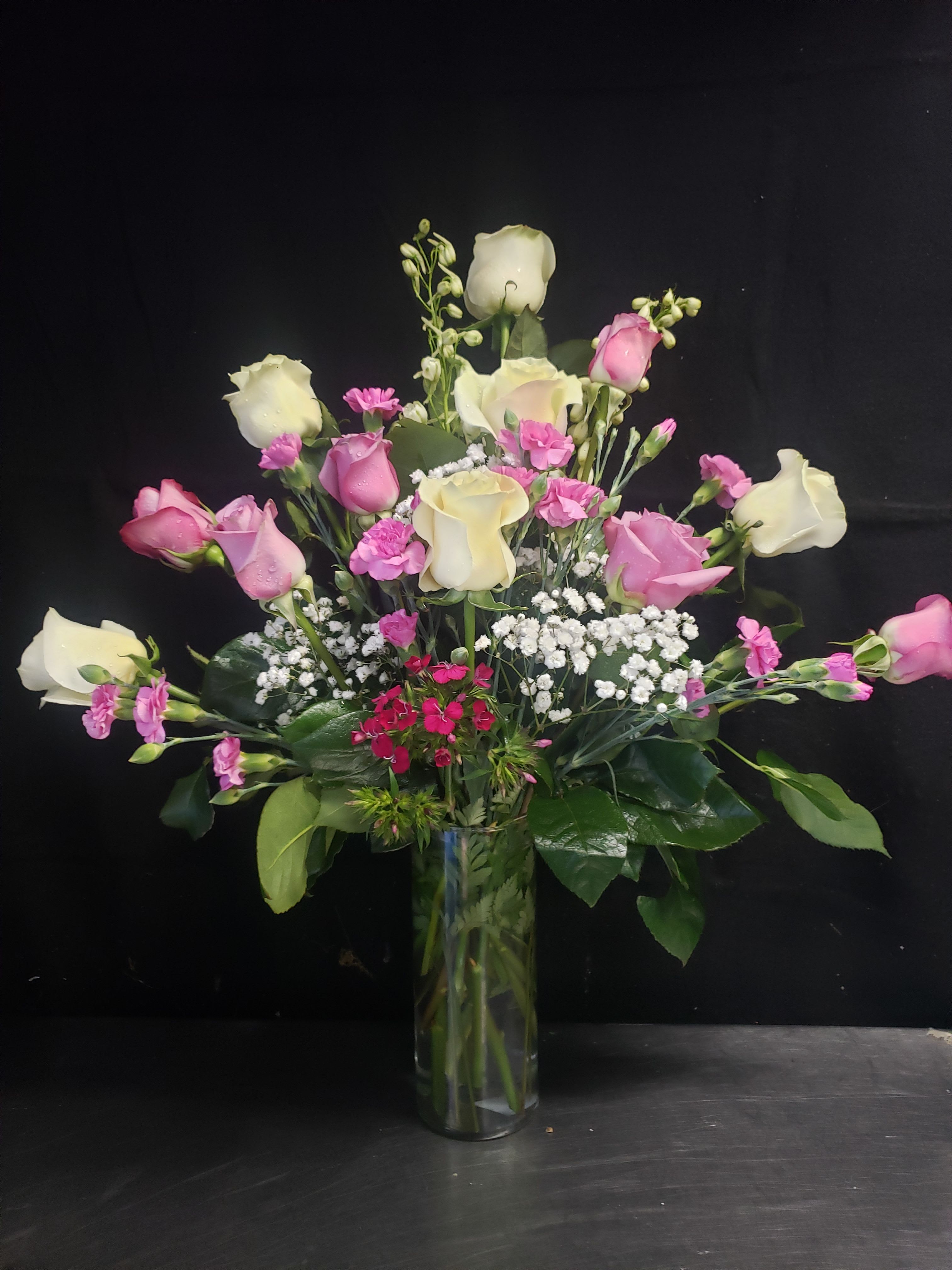 I love Pink  - Soft blush and vivid pink roses reach upward in a tall clear glass vase, making this flowers for events arrangement perfect for anniversaries, birthdays, or a wedding reception. The airy, asymmetrical silhouette-rose heads extending outward with pops of bright pink spray blooms-reads romantic and celebratory, the kind of bouquet that makes the person you're thinking of feel seen and adored.  Alongside the roses are small pink carnation-like blooms and clusters of tiny white filler flowers (baby's breath) woven through glossy dark green foliage for texture and contrast. The palette of pink, white, cream, and green gives a cheerful yet elegant impression; the clear glass vase keeps the focus on the open, full shape and lets stems show for a fresh, modern look.  Ideal for table accents, ceremony décor, or a thoughtful gift, this arrangement arrives ready to display and suits both casual everyday gifting and formal events. For best longevity, refresh the water every 2-3 days and trim stems at an angle when you change the water to keep the roses and fillers looking their freshest.