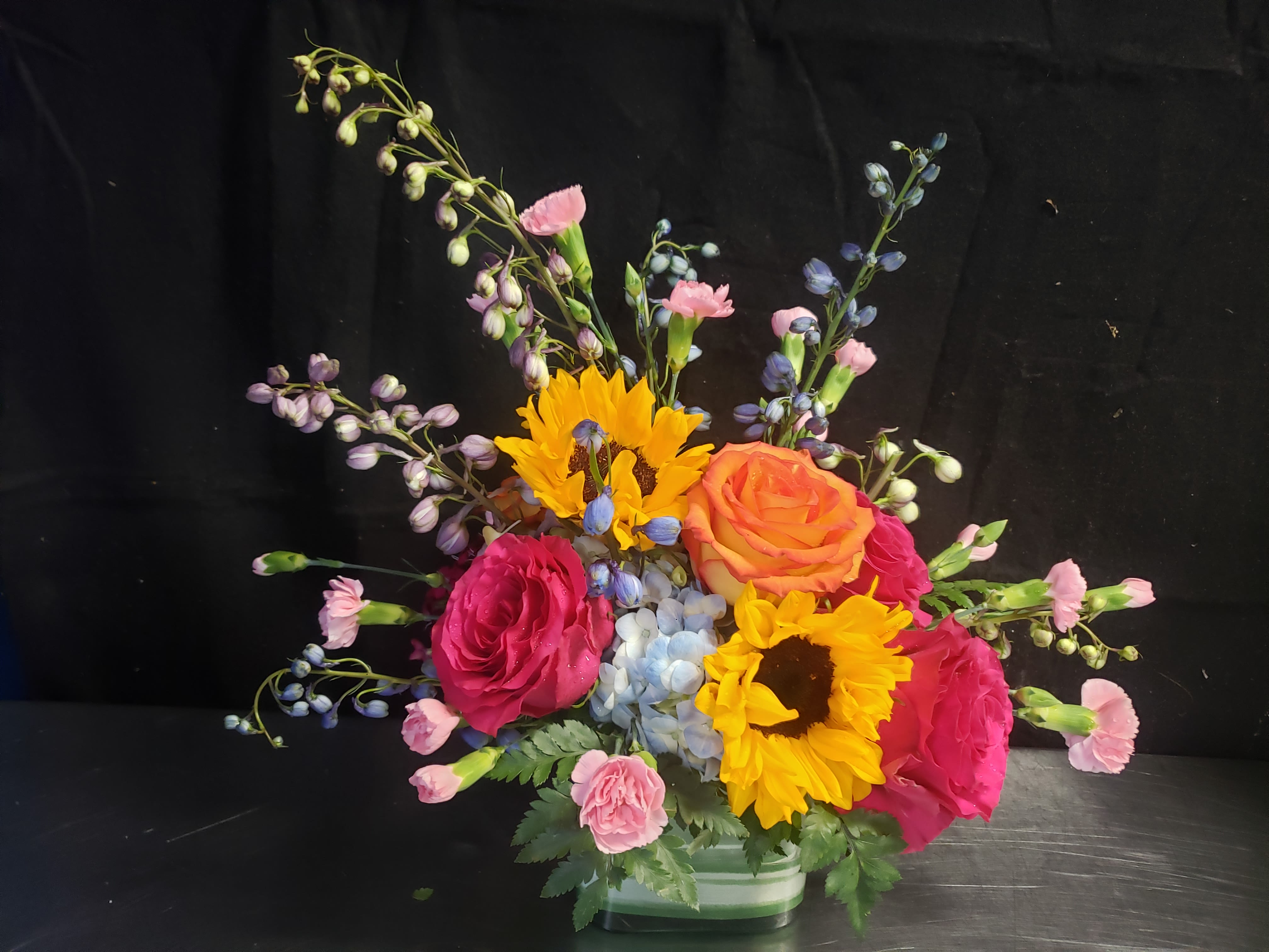 CIDIANT CITRUS BOX - A sunny burst of color that reads like a celebration in bloom - this cheerful flower arrangement is perfect as a birthday bouquet, congratulations gift, or an energetic just-because surprise. Pink and orange roses set a warm, affectionate tone while bright yellow sunflowers bring instant joy; the mix signals celebration and makes the recipient feel genuinely seen and celebrated the moment they walk in.  Pale blue hydrangea forms a soft, cool focal at the center, surrounded by delicate pink carnations and airy blue and lavender filler stems that fan outward for a loose, garden-style silhouette. The composition balances compact weight at the base with upward movement from taller stems, creating a lively, textured look. Available in a low glass vase or a low ceramic vase, this arrangement highlights mixed blooms - roses, sunflowers, hydrangeas, carnations, and seasonal accents - in pink, yellow, orange, blue, and purple tones.  Ideal for sending to a friend, colleague, or family member to mark milestone moments or brighten an ordinary day, the design arrives ready to display on a table or mantle. The low vase keeps it easy to place and admire; simple fresh-water care (change water and trim stems every few days) will help the flowers stay vibrant while they do the work of making someone's day.