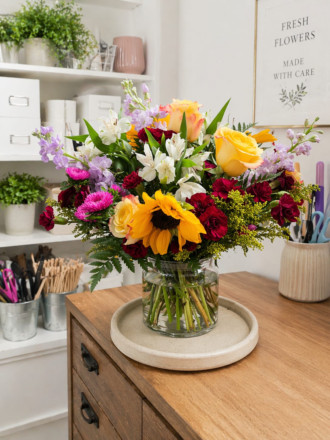 Garden delight  - A vibrant, hand-tied arrangement featuring sunflowers, yellow roses, red spray roses, lavender stock, and white alstroemeria, designed in a clear glass vase for a bright, cheerful, and elegant look. Perfect for birthdays, celebrations, or everyday joy.
