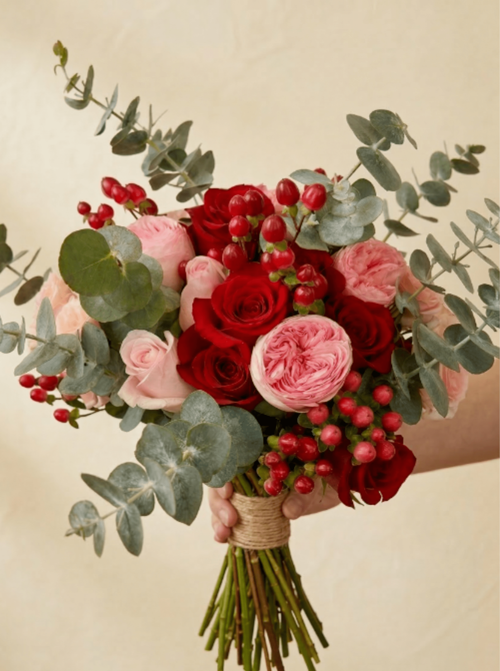 Hand-tied bouquet of red and pink roses with red berries and eucalyptus, held against a beige background.
