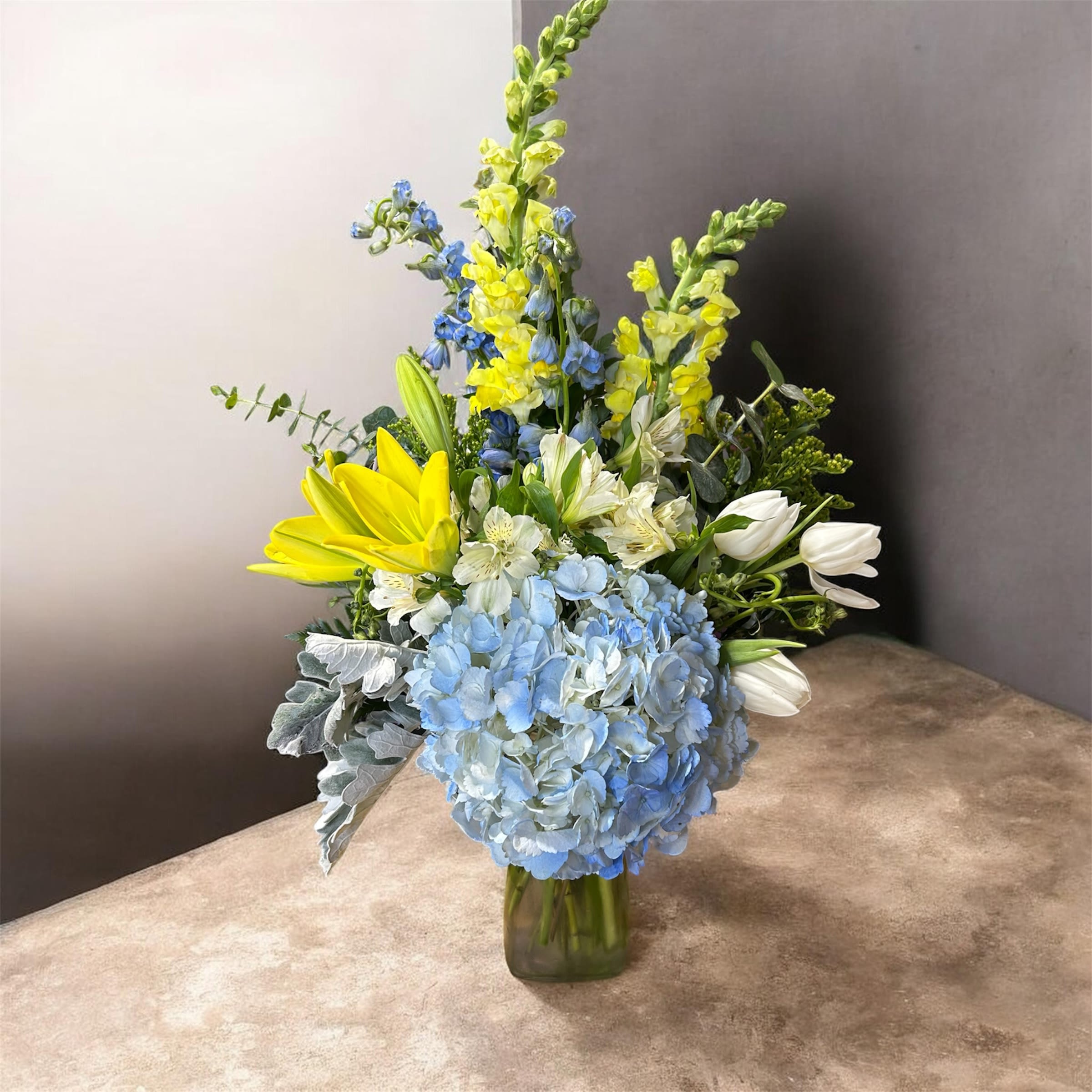 Joyful Bouquet  - Pale blue hydrangeas form a soft, cloudlike focal point in this spring bouquet, surrounded by tall yellow and blue snapdragons that give the arrangement a graceful, vertical silhouette-perfect as a cheerful birthday bouquet, thank-you gift, or everyday celebration. White alstroemeria and white tulips tuck in around the center and right side, their crisp petals brightening the cool-blue and green tones so the recipient feels both uplifted and thoughtfully noticed.  Arranged in a clear glass vase, the mix of textures-from the hydrangea's lush mound to the snapdragons' spiky stems and the tulips' smooth cups-creates an airy, slightly formal presentation that still reads fresh and approachable. Bouquet height and vase size make it an easy centerpiece for a dining table or an elegant desk accent; refresh stems and change water every few days to keep blooms looking their best.