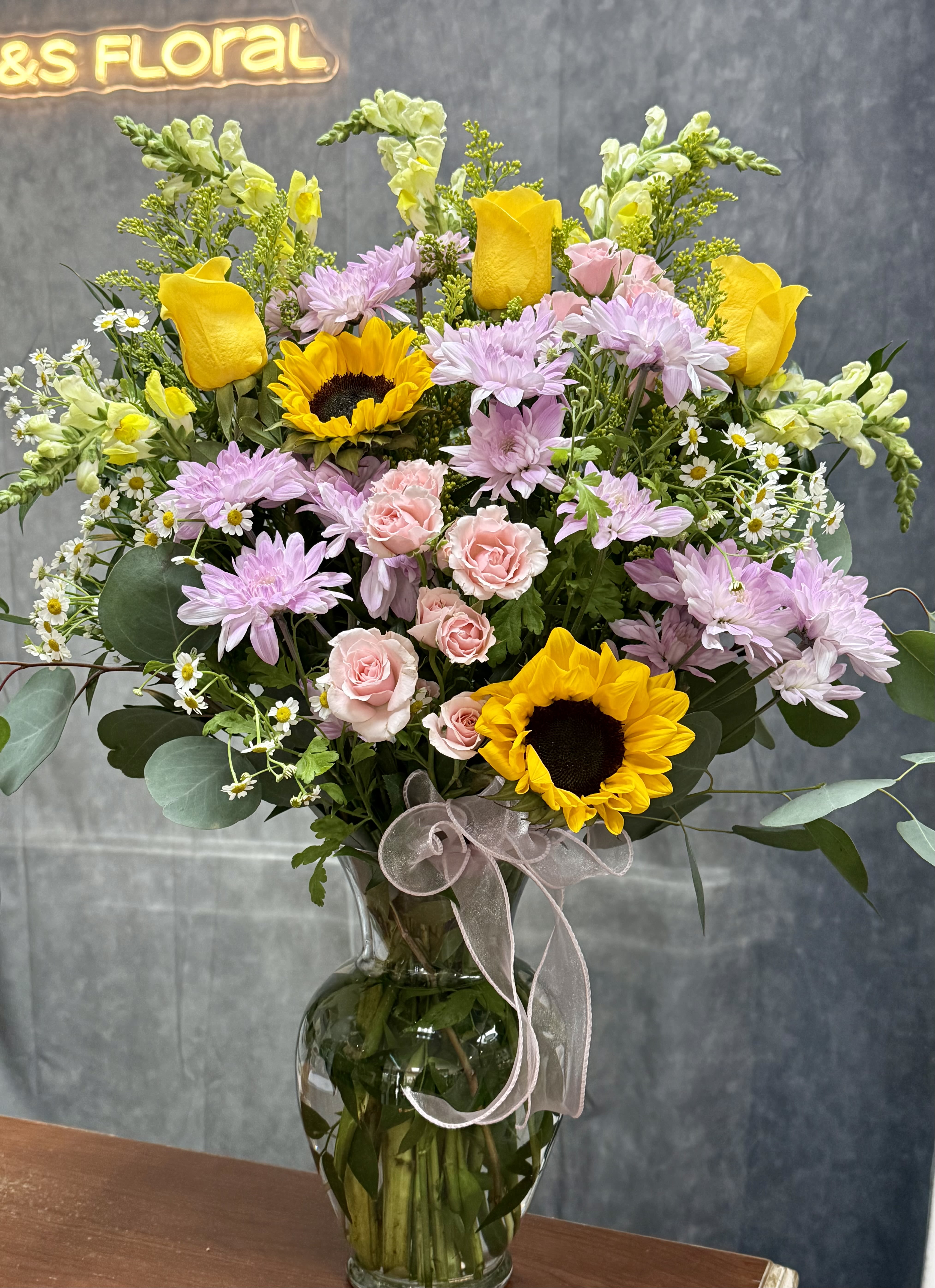 Garden Sunrise Vase - A radiant mix of golden sunflowers, sunny yellow roses, delicate pink spray roses, and cheerful lavender daisies — a vibrant vase arrangement that feels like a warm morning stroll through a blooming garden.