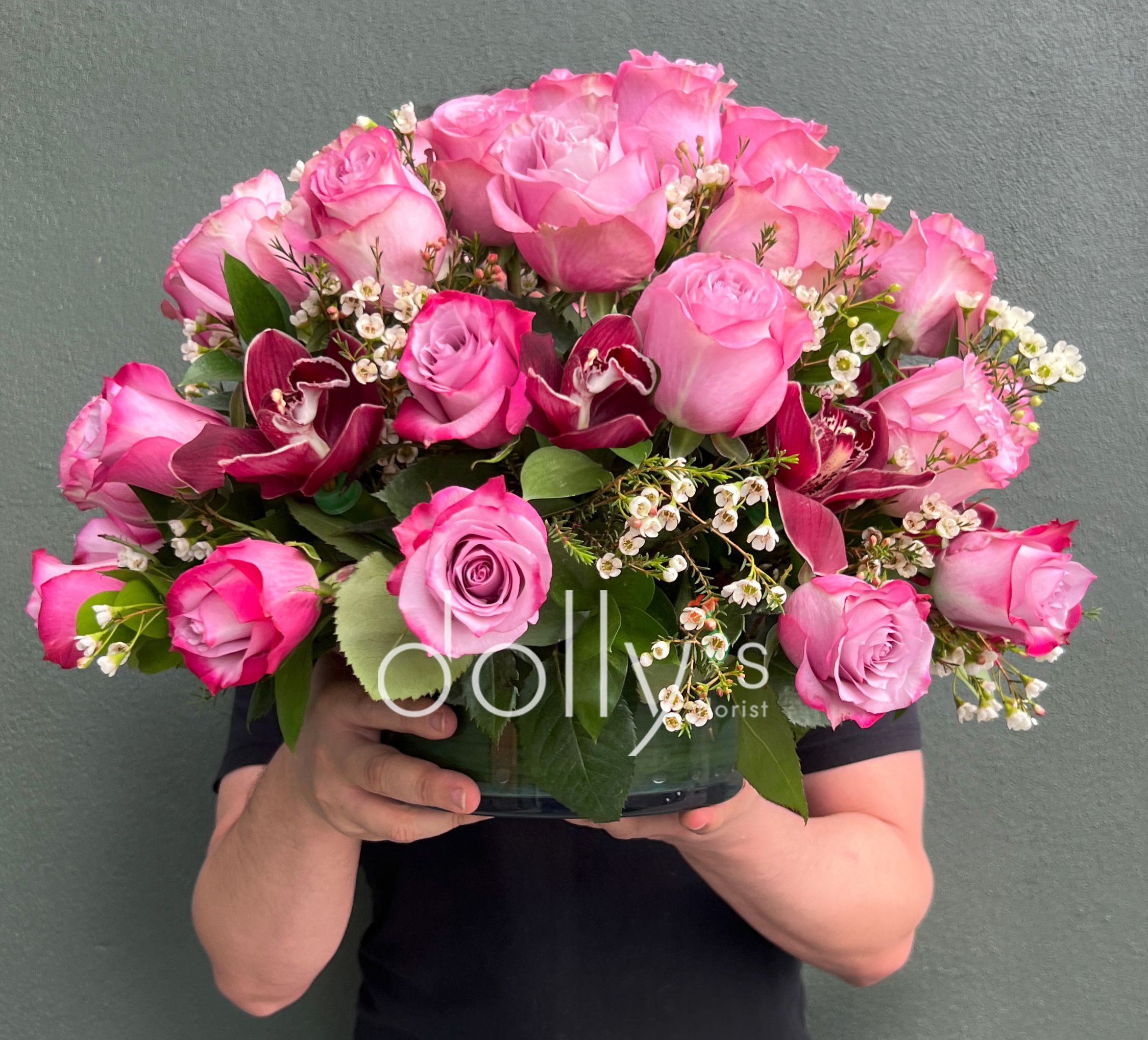 Blushing Velvet - A lush dome of blush and hot-pink roses crowned with deep burgundy cymbidium orchids creates an immediate romantic focal point-perfect as an anniversary bouquet, birthday arrangement, or a Mother's Day gift that makes her feel truly celebrated. This compact flower arrangement also reads beautifully for love and romance, thinking of you, thank you, get well, or I'm sorry moments when you want the recipient to feel cherished.  Carefully gathered into a low bowl, the design pairs 36 premium pink roses with exotic orchids and small white filler blooms for contrast and texture. The rounded, low-profile silhouette sits nicely on a desk or tabletop and brings rich color without overwhelming a space; the orchids' patterned centers add visual depth while the white accents keep the palette fresh.  Choose this arrangement when you want someone to feel noticed-whether congratulating a milestone, marking Valentine's Day, supporting Breast Cancer Awareness Month, or simply saying you care. Arrives ready to display; keep in fresh water, out of direct sunlight, and enjoy the blooms for days as you refresh the vase water and trim stems every few days.