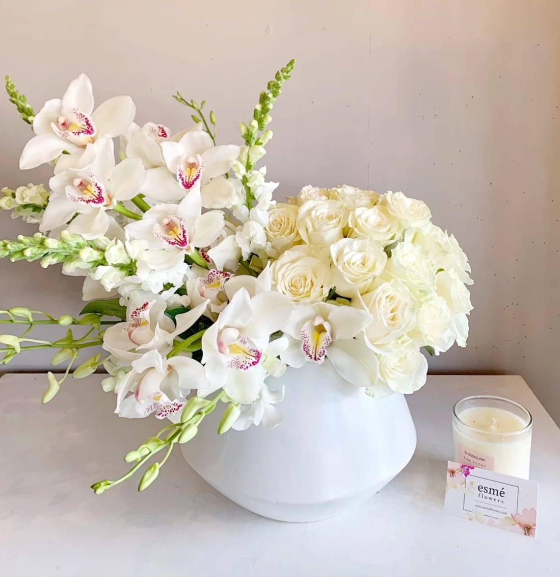 White roses and orchids arranged in a white vase beside a candle.