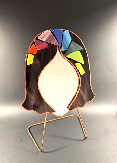 #A026 - Burgundy Mosaic Face Mirror #2 - A playful stained-glass art mirror that feels like a tiny celebration - accented decorative piece is perfect as a Mother's Day gift or a vibrant birthday or housewarming surprise. The arrangement pairs mixed-color mosaic accents with white, blue, and yellow tones around a smooth, light-colored central face, creating an uplifting focal point that reads as both modern sculpture and functional mirror. Its abstract human form and bright geometric tiles make the recipient feel seen and celebrated-great for anniversaries, graduations, get-well cheer, or just because.   Set on a slender metal stand, the stained-glass mirror combines darker marbled glass framing the face with a playful crown of red, orange, yellow, pink, blue, and green mosaic pieces along the top edge. The piece averages about 11½" in height and is designed to sit neatly on a shelf, desk, or side table; each mirror's mosaic pattern and overall height vary slightly. Special orders are welcome-message us with requests-and domestic shipping within the contiguous U.S. is free.  Shipping available - select Delivery, then message us to ship. 