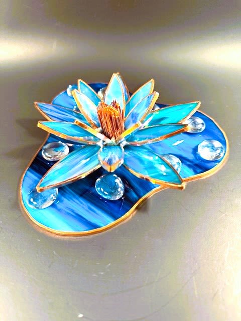 #A028 - Azure Iridized Water Lily - A marbled blue-and-teal stained-glass lotus brings a quiet, reflective moment to any room-perfect as a tabletop sculpture for a birthday, housewarming, or sympathy gift. The piece reads like a floral sculpture: layered, pointed petals rise toward a textured, copper-toned center so the person you're thinking of will feel seen and comforted by its calm, iridescent glow. Handcrafted from marbled blue, teal, and white stained glass with warm metallic edging, the lotus sits on a matching lily-pad base and is finished with clear glass pebbles and gems that catch the light like water droplets.   The compact, dimensional design works beautifully on a desk, shelf, windowsill, or mantel as a lasting keepsake that complements fresh flowers or stands alone as an artistic accent. Give this as a thoughtful token to mark quiet occasions-sympathy, reflection, a calming birthday gift, or simply to say you care-and choose it when you want a present that endures. The sculpture's size and sturdy hard goods make it easy to place anywhere; wipe clean gently with a soft cloth to preserve the glass and metallic highlights.  Shipping available - select Delivery, then message us to ship.