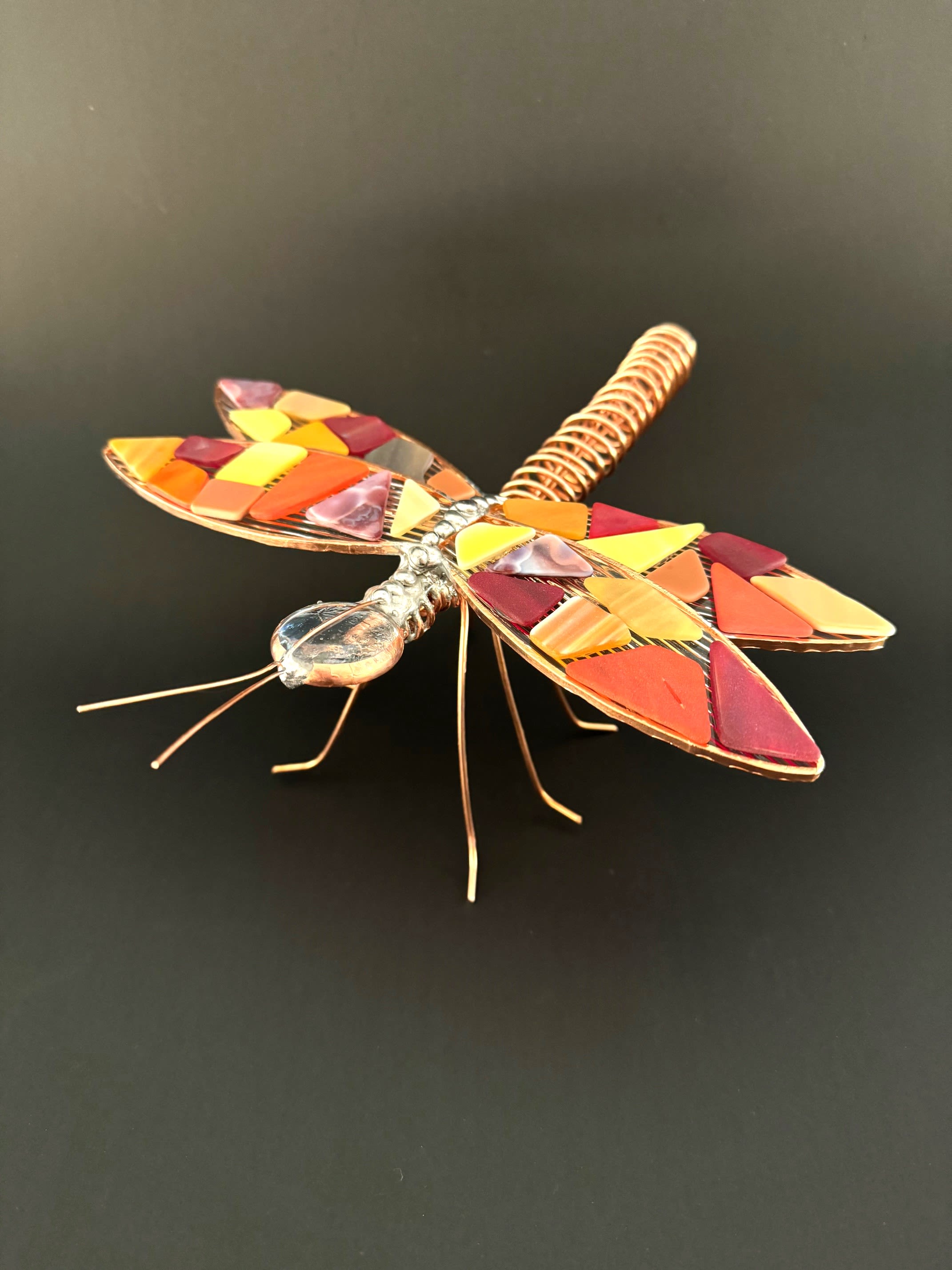 #A036 - "Fire" Mosaic Dragonfly - A playful, handcrafted dragonfly accent that brings warm, autumnal color to any tabletop or bouquet - perfect as a seasonal gift accent or a whimsical housewarming touch. This decorative metal and mosaic glass sculpture works beautifully as an accent for an anniversary bouquet or as a standalone home décor piece to celebrate someone who loves nature and crafty details.   The piece features a coiled, copper-toned metal body with fine wire legs and antennae for a delicate, handmade profile. Its wings and inlay are filled with small mosaic glass tiles in shades of orange, yellow, red, pink and maroon, creating a jewel like effect that glows against darker backgrounds. The contrast of shiny metal and matte mosaic gives it an artistic, tactile quality. Tuck it into a mixed-color arrangement to add sparkle and texture, set it beside a potted plant, or present it on its own as a thoughtful accent that says you notice the little things. Lightweight and easy to place, it's ideal for seasonal décor, hostess gifts, or a charming finishing touch to a gift bouquet.  Shipping available - select Delivery, then message us to ship.