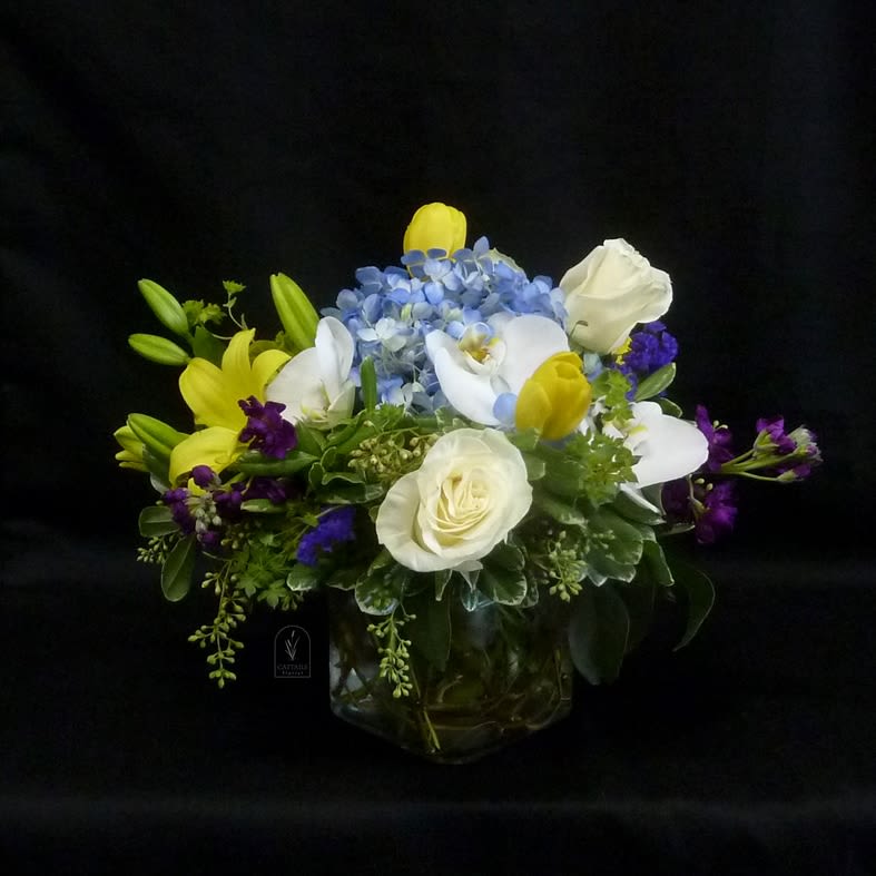 Age of Aquarius - Age of Aquarius is a fresh, uplifting compact arrangement with soft whites, sunny yellows, and calming blues. Featuring elegant roses, cheerful tulips &amp; lilies, delicate orchids, and lush hydrangea, it creates a harmonious balance of light and serenity.