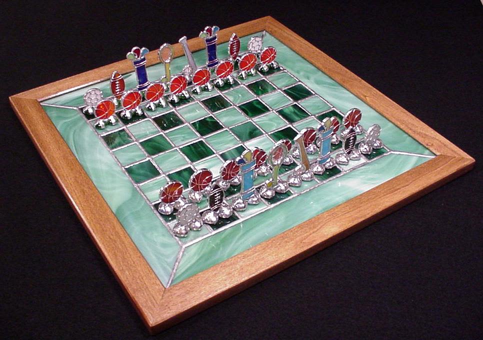 #A089 - Sports Chess Set - A jewel-like decorative chess set that reads as art as much as a game - perfect as a gift for a sports fan, a game-room accent, or a thoughtful birthday or housewarming present. This is a stained-glass chess board set in a warm wooden frame, making an immediate visual statement for an office, living room, or den where you want to say "I notice the details" to the person you're buying for.   The board's playing surface alternates translucent and darker green glass squares divided by silvery metal grid lines, giving a luminous, faceted look that catches light like a small mosaic. Chess pieces are metal and glass sculptures-polished metal bodies with red, blue, white and black glass accents and tiny sports motifs that add playful, collectible character while remaining elegant on display. The mix of warm wood, cool metal, and colored glass creates a tactile contrast that reads as both modern and handcrafted. This set works as both display and play: it's the kind of gift that makes the recipient feel seen for their hobbies and taste, whether marking a special occasion or simply saying "I was thinking of you." Its decorative quality suits shelving, a side table, or a game nook; handle gently and avoid direct impact to the glass pieces when playing or moving the set.  Shipping available - select Delivery, then message us to ship. 