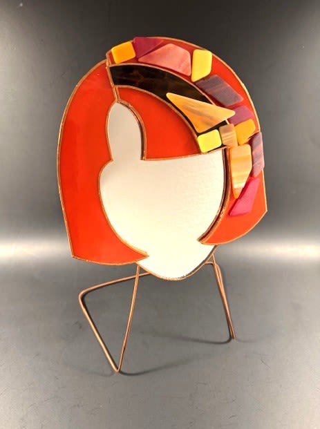 #A091 - Bright Orange Mosaic Face Mirror #2 - A bold stained-glass silhouette that reads like a joyful conversation piece, this bright orange mosaic face mirror makes a memorable gift for Mother's Day, birthdays, anniversaries, or as a special housewarming accent. This quirky mirror is made with artful design - think of it as a floral-forward keepsake that says "I'm celebrating you" whether the occasion is a wedding, retirement, graduation, or just because. The piece features a modern head-shaped panel in saturated orange with a mirrored facial panel framed by a band of small geometric mosaic tiles in yellow, red, purple, and amber tones.   Mounted on a minimal metal display stand with thin angled legs, the sculpture has a light, sculptural presence that complements living rooms, entryways, or office desks. The warm palette reads sunny and uplifting-perfect alongside spring and summer blooms or as a standalone accent to brighten a bedside table or mantel. Give this to the person you're thinking of when you want them to feel seen and celebrated: it works beautifully for love and romance, thank-you gifts, get-well wishes, or to mark holidays from Christmas to Hanukkah. Its compact, freestanding design makes it easy to place.  Shipping available - select Delivery, then message us to ship.