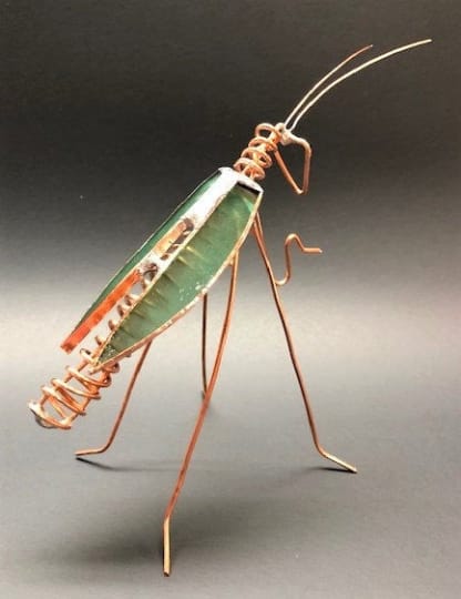 Metal wire grasshopper sculpture with a green body on a dark background