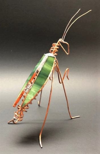 #A105 - Ivy Green Praying Mantis - A whimsical glass-and-wire insect that delights at first glance, this small sculptural piece makes a playful gift for birthdays, house warmings, or someone who loves unique desk accents. The modern-style sculpture reads like a miniature art object-a green-themed accent with a slim stripe of copper surrounds each wing and creative wirework will make the person you're thinking of smile and invite compliments. Handcrafted from slender copper-toned metal wire and a green stained-glass body, the piece features long delicate legs, a curled torso, and extended antennae that create an airy, sculptural silhouette.   The stained glass shows variegated green tones with a narrow orange edge for contrast, and the metal wire has a dark, almost black finish in places that adds a modern, slightly industrial touch. Compact enough for a shelf, windowsill, or among potted plants, it brings texture and a touch of handmade charm without overwhelming a space. Perfect as a casual gift to celebrate a new home, a birthday, or to brighten a workspace, the piece arrives ready to display-no special care required beyond gentle dusting. Its small scale and durable materials make it easy to place wherever this little mantis will catch the light and the recipient's eye.  Shipping available - select Delivery, then message us to ship.