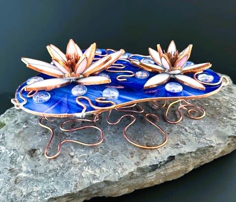 #A126 - White and Pink Floating Water Lilies - Soft white and blush stained-glass lotus blooms float above a vivid blue glass "pond," creating a tranquil sculpture, perfect as a unique decoration for Mother's Day, an anniversary, or a thoughtful housewarming gift. The floating water lilies read like a peaceful centerpiece that will make the person you're thinking of feel seen and soothed - an elegant choice for birthdays, get-well wishes, retirement celebrations, or simply "just because."   Two stylized lotus flowers in white and pink, edged in warm copper, rest on an oval blue glass base scattered with clear glass pebbles that catch the light like water droplets. Curving copper wire forms a looping pedestal that lifts the blooms above the surface. Size and materials give it lasting presence on a mantel, bedside table, or entry console.   This keepsake-style arrangement works year-round-from Mother's Day and summer celebrations to holiday gifting, graduations, and weddings-offering low-maintenance beauty without floral care. It's an especially meaningful pick for someone who appreciates artful décor and quiet moments; choose it when you want a gift that conveys calm, gratitude, or heartfelt apology.  Shipping available - select Delivery, then message us to ship.