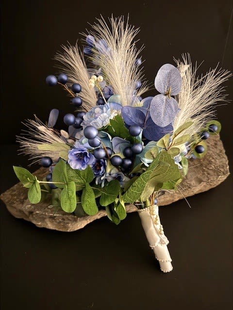 #A118 - Blue Hydrangea Silk Floral Bouquet - Soft blue hydrangeas and velvety ranunculas form the heart of this silk wedding bouquet, arranged with delphinium accents and jewel-toned navy berry clusters for depth. Designed for weddings, vow renewals, or anniversary celebrations, the slightly asymmetrical hand-tied shape feels modern and textural-more sculptural than tightly domed-so the person you're gifting will look and feel effortlessly refined and seen.   Silk blooms are paired with feathery ivory plumes and muted green-blue foliage to create contrast in both color and texture; smooth petals sit against matte berries and spiky dried accents for a layered, tactile effect. The stems are finished with an ivory ribbon-wrapped handle and delicate pearl details for a bridal-ready hold.   Available as a handheld bouquet or as a centerpiece option gathered in a clear ribbed glass vase (or presented on a stone display slab), this arrangement suits romantic, natural, or winter-inspired weddings and keeps its cool blue-and-white palette beautiful year-round. Care: silk construction means it arrives ready to wear or display with no wilting-spot clean dust as needed.  Shipping available - select Delivery, then message us to ship.