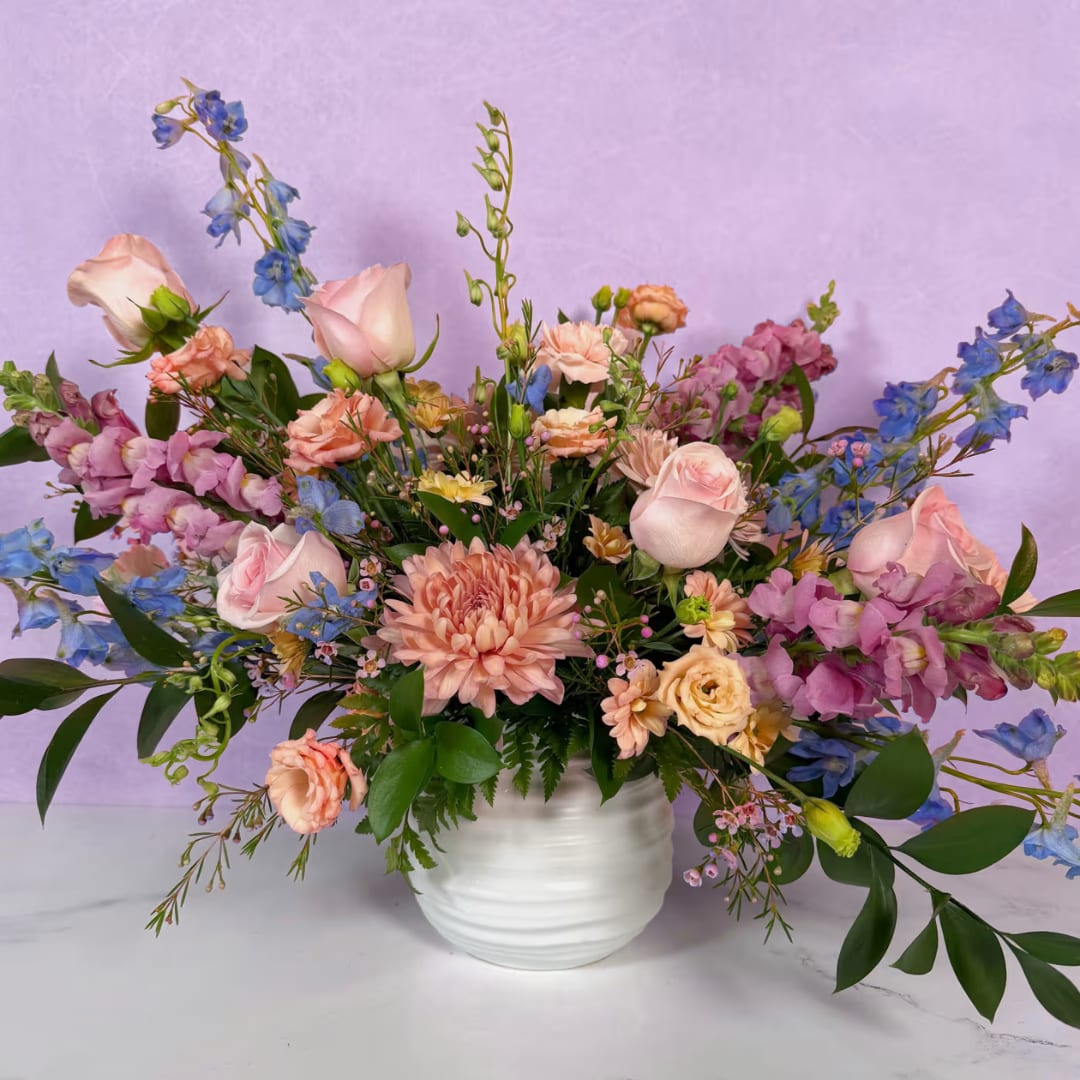 English Garden - She'll be the talk of the town when you surprise her with her very own English Garden! This design looks right out of a royal garden, with beautiful pastel blooms like blue delphinium, light pink roses, blush cremones, and more gracing this exquisite design. All of these decadent flowers are expertly crafted in a stylish white ceramic larkin pot, and the whole thing comes together to create a magnificent display fit for royalty!  Approx. L 27" x H 22"