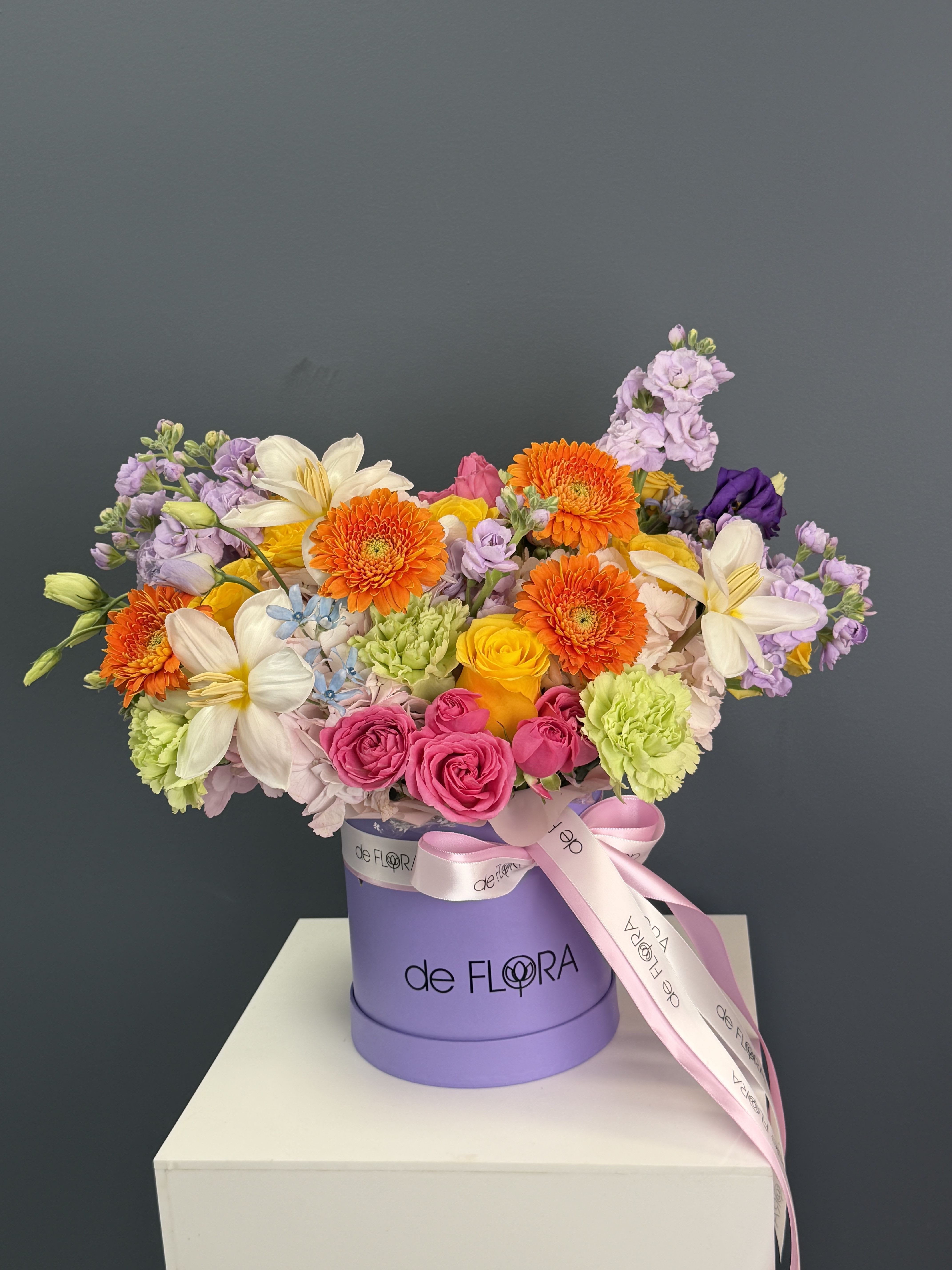 Sunset Bloom Box - A vibrant, garden-inspired composition designed to impress at first glance. This elegant arrangement features a playful mix of premium blooms — radiant orange gerberas, delicate lilies, soft lavender stock, fresh green carnations, and roses in shades of blush pink and golden yellow. Accented with airy textures and seasonal florals, it creates a perfect balance between brightness and softness.  Presented in a signature deFLORA hat box with a satin ribbon, this arrangement feels both modern and joyful — ideal for birthdays, celebrations, or simply bringing color into someone’s day.