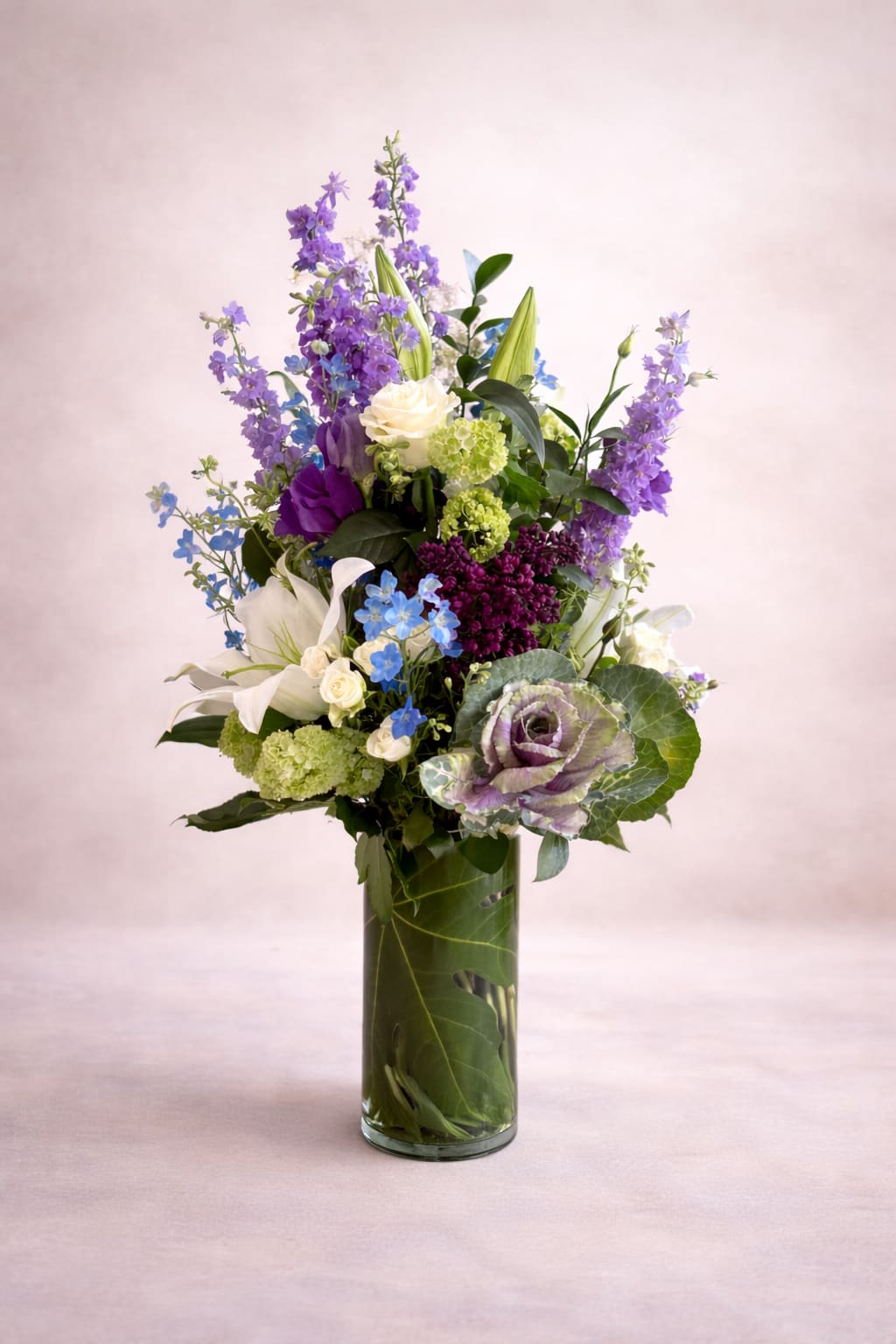 English Garden - A tall, airy garden-style arrangement rises gracefully from a clear cylinder vase lined with a broad green leaf, creating a clean, modern base for the abundant florals above. Soft ivory roses and creamy white lilies anchor the composition, while vertical spires of lavender delphinium add height and movement. Clusters of vibrant green hydrangea provide fullness, complemented by delicate blue delphinium accents and a rich band of deep purple lilac near the center. A striking ornamental cabbage bloom introduces a sculptural, textural focal point, while layered greenery and varied blooms create a lush, natural, and elegantly unstructured silhouette.