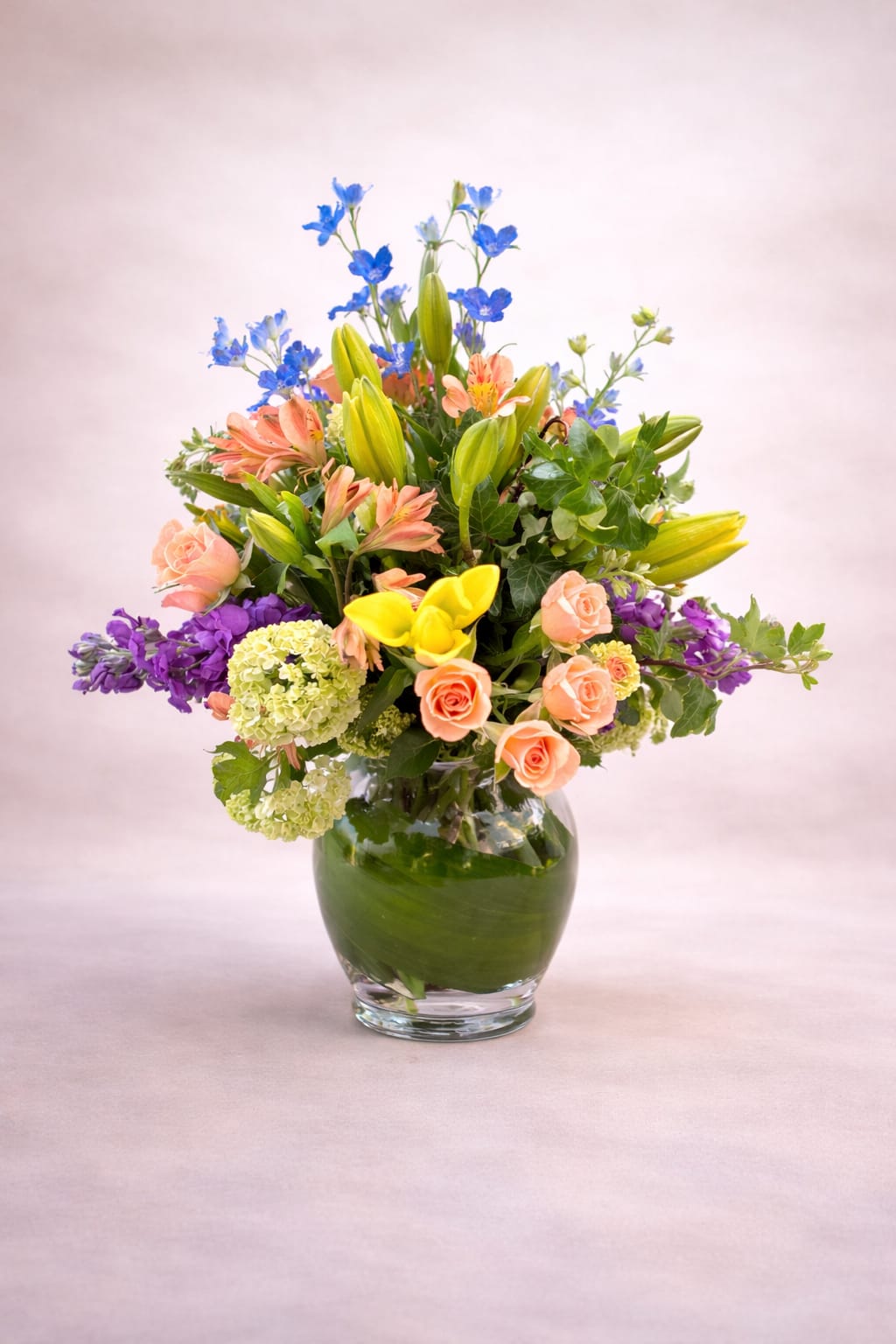 Secret Garden - This bouquet feels lush and garden-fresh, composed with a vibrant mix of color and texture that creates a lively, layered effect. Upright lily buds and airy stems of soft blue flowers add height and movement, while clusters of peach roses bring warmth and softness throughout the arrangement. Bright yellow blooms offer cheerful focal points near the center, balanced by pale green hydrangea that ground the design with fullness. Accents of purple flowers and varied greenery weave through the composition, all arranged in a rounded glass vase lined with rich green leaves for a polished, natural finish.