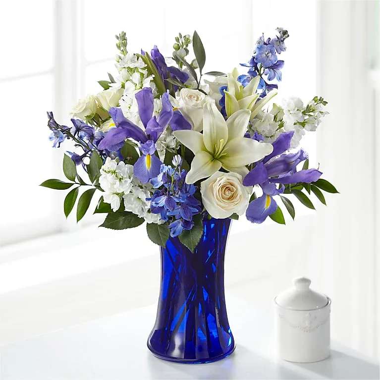 Blue Axis - A cool, airy bouquet of white lilies and roses with vivid blue delphinium and purple irises-this flower arrangement is an elegant everyday gift and makes a refined birthday, thank-you, anniversary, or sympathy arrangement. The tall cobalt-blue glass vase gives the composition a polished, formal contrast that lets the crisp whites and saturated violets sing; the overall mood is serene with a touch of romance the recipient will notice at first glance.  The design is full and rounded, layered with large white lilies, clustered white roses, spikes of blue delphinium, and purple irises, plus small white filler blooms and verdant greenery for texture. Arranged in a tall cobalt-blue glass vase (also shown in a ceramic jar option), the piece reads like a garden-style composition with airy vertical accents and a lush, dense crown of flowers. Sized for tabletops or entryway display, this is a thoughtful way to show someone you're thinking of them-order for next-day delivery where available and follow simple care: fresh water and a cool spot will keep the blooms looking their best.