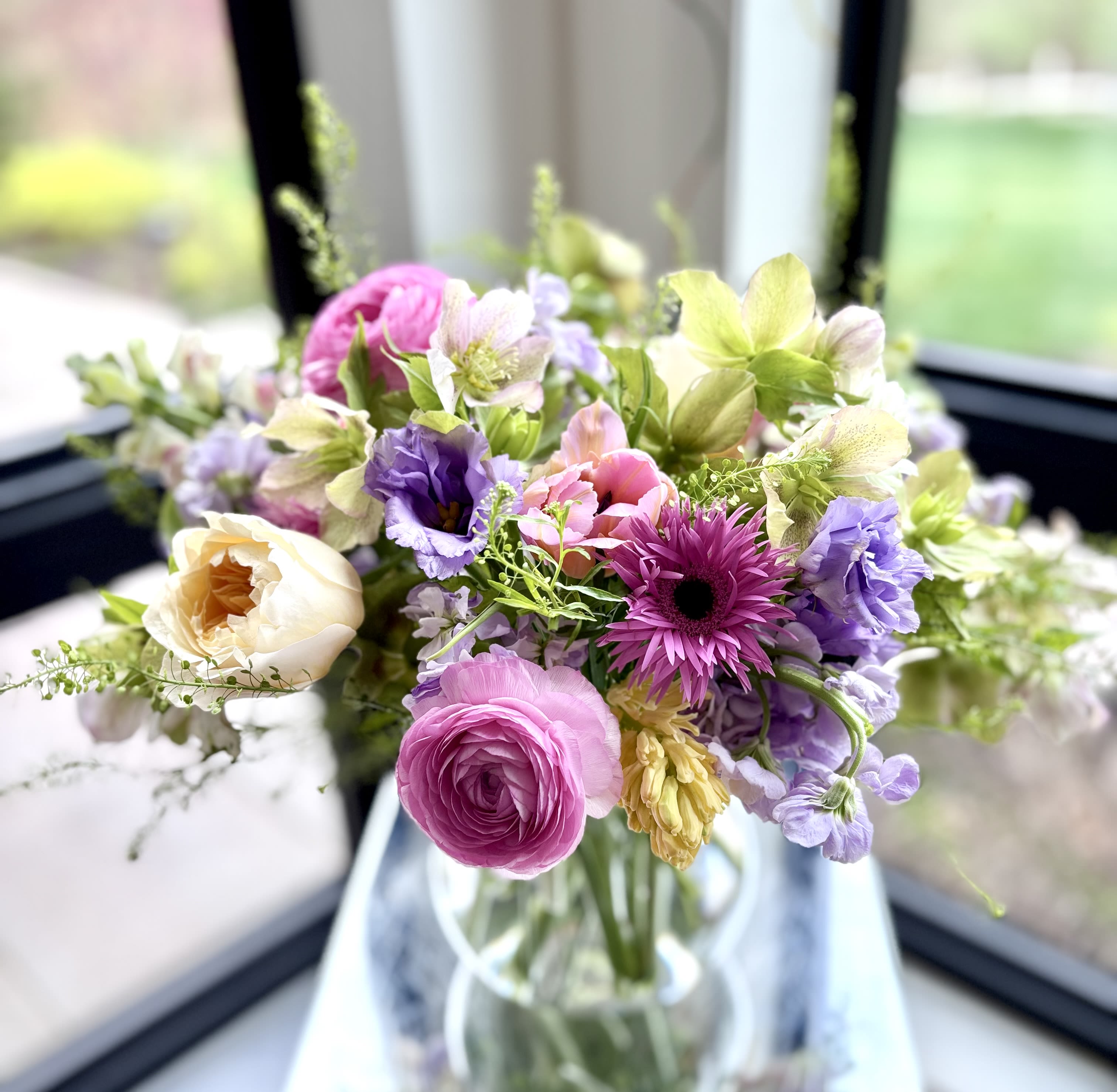 Violet - A lovely fresh flower arrangement in Lula vase with lovely blooms such as garden rose, hellebore, anemone, tulips, snapdragons, stock, lisianthus, gerbers and additional fillers based on the availability of flowers, in the color palette of yellows, pinks, lavenders, greens and whites.