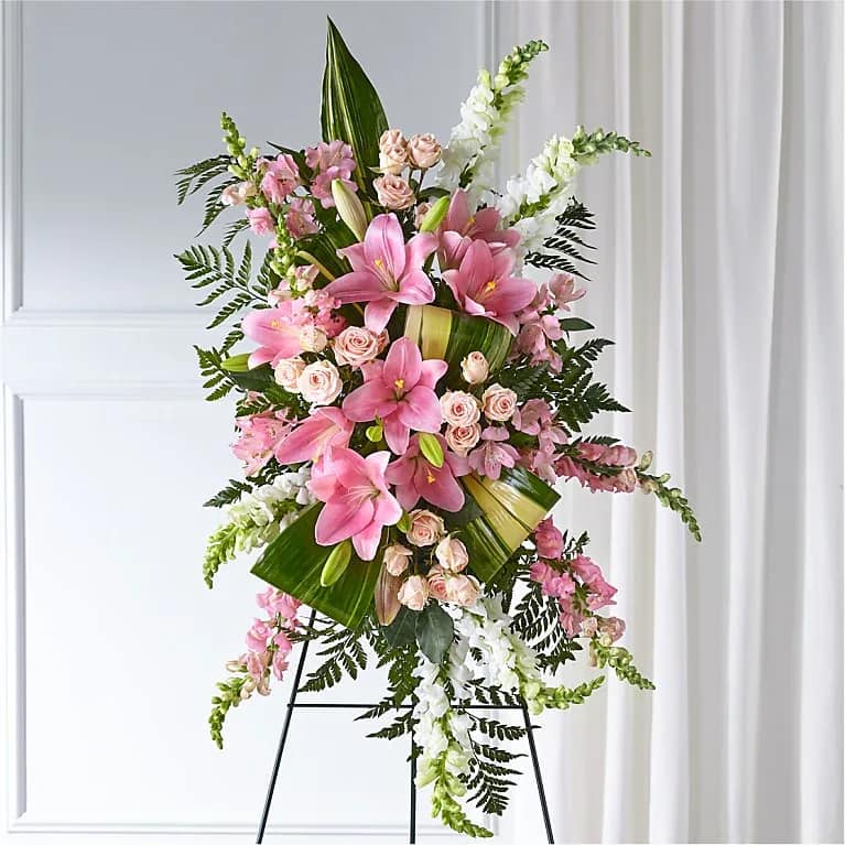 Florent Veil - A tall, ceremonial standing arrangement that reads as gentle and romantic-perfect as a sympathy spray or a formal celebration piece. This flower arrangement for memorials or commemorations features large pink lilies and pale pink spray roses framed by white snapdragon spikes and fresh alstroemeria, offering a graceful, uplifting presence that will make the person you're honoring feel remembered and seen.  Broad tropical leaves and fern-like foliage build a layered vertical structure while cascading blooms soften the silhouette, blending pink, white, and green for a soothing palette. Mounted on a black metal easel for a display-ready presentation, the arrangement's height and formal lines make it easy to place at a service or event; approximate dimensions and care instructions are available at checkout to help you choose the right size and ensure flowers stay fresh through the occasion.