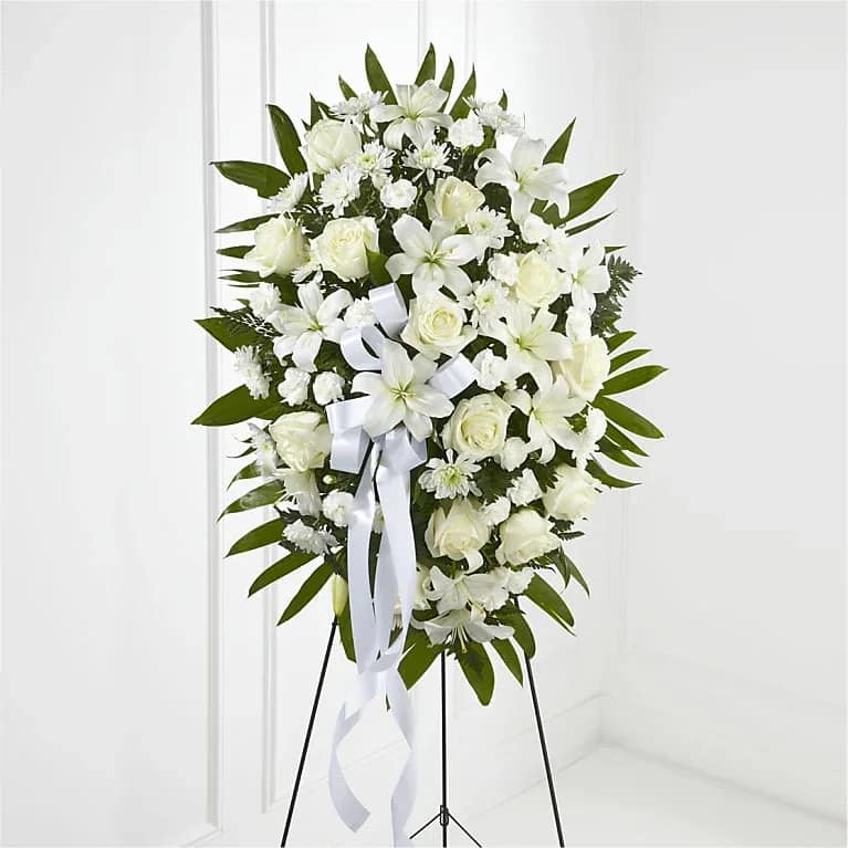 Lumen Wreath - A large white standing spray arranged in a classic oval-this sympathy flower arrangement combines white roses, lilies, daisies and chrysanthemum accents to create a calm, respectful tribute for a funeral or memorial service. The all-white palette and formal silhouette send a quiet, comforting message to the person you're honoring, offering a dignified way to say you're thinking of them.  Flowers are densely clustered into an oval shape and framed with broad dark green foliage for high contrast and structure. A long white ribbon cascades from the center, and the spray is mounted on a black metal standing easel for prominent display. Sized as a large standing spray, it's designed for traditional services and graveside presentations; minimal care is required beyond keeping the stems hydrated until placement.