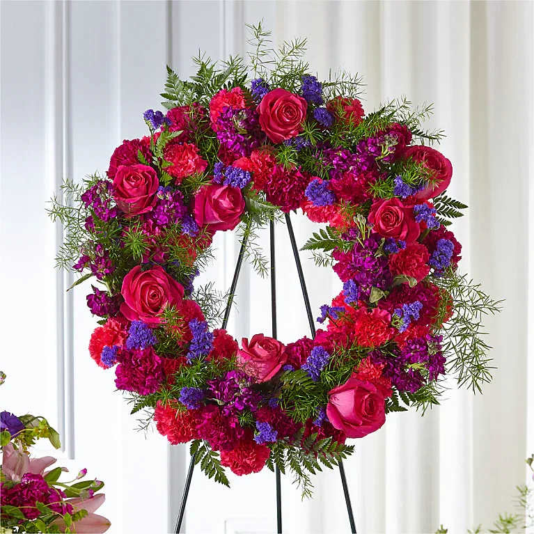 The Shape of Heat - A bold heart-shaped wreath that speaks of love and remembrance-the Shape of Heat is a heart funeral or sympathy arrangement made to honor someone special or mark a deeply romantic moment. Bright pink roses and clustered red and magenta carnations create the dominant heart, while touches of small purple filler blooms and airy fern-like greenery soften the edges and add texture. Mounted on a black metal easel stand, this piece is designed for a front-facing, formal presentation that commands attention.  This wreath's saturated pinks, reds, and purples read warm and celebratory while remaining respectful, making it appropriate for memorial services, sympathy displays, or a striking anniversary or Valentine's tribute when a larger, more formal statement is wanted. The dense, textured composition gives visual depth from a distance; the metal easel provides sturdy, upright display. For placement, plan to present it where it can be viewed head-on so the heart shape and layered florals make the strongest impression.