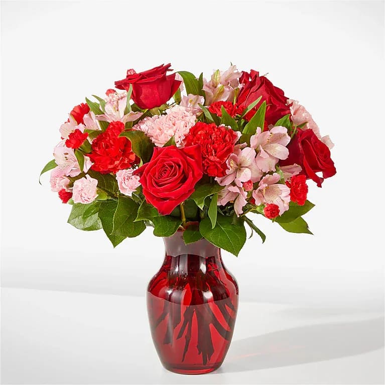 Well Contained - A lush cluster of red roses and pink carnations gathered tight in a glossy red glass vase-this flower arrangement is a cheerful, romantic way to celebrate an anniversary, birthday, or any love-and-romance moment. The rounded, full silhouette reads classic and substantial, the saturated reds paired with pale pinks giving a bold yet tender visual that will make the person you're thinking of feel truly seen.  Packed closely for a dense, garden-fresh look, the bouquet blends red roses, red and pale pink carnations, and light pink alstroemeria for contrast and texture; glossy green foliage peeks through to lift the palette. The arrangement arrives in a coordinated red glass vase (also shown in a clear glass option), creating a polished presentation that suits both everyday gifting and special occasions. Sized to sit beautifully on a dining table or desk, it offers long-lasting color-order with same-day delivery where available to make a timely, heartfelt impression.
