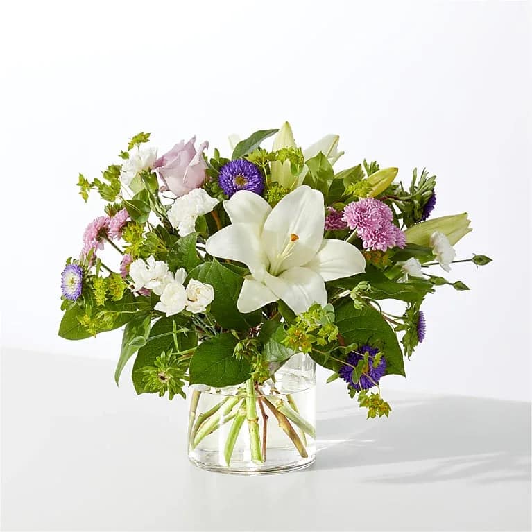In a Balanced Quiet - White lilies take center stage in this airy garden-style vase arrangement, a gentle everyday bouquet perfect for birthdays, anniversaries, sympathy, or a thinking-of-you surprise. The fresh white focal blooms paired with soft lavender roses and pink chrysanthemums create a calm, romantic mood that will make the person you're thinking of feel comforted and truly seen.  The loose, rounded design mixes white lilies, pale lavender roses, pink chrysanthemum pompons, purple daisy-like asters and small white filler flowers with airy greenery, all gathered in a clear glass vase. Blossoms are layered at varied heights for a natural, slightly wild look that reads both elegant and relaxed. This arrangement arrives ready to display; swap the water and trim stems every few days to keep blooms looking their best.