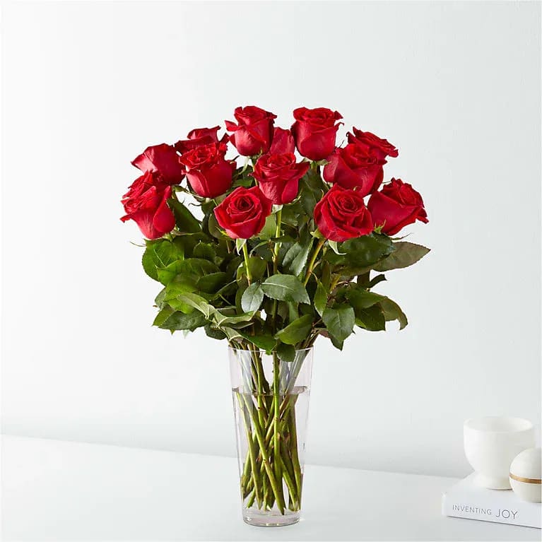 True, Untouched - A dozen red roses gathered in a tall clear glass vase creates a classic, romantic statement-the kind of bouquet that makes the person you love feel cherished. Perfect for a birthday, anniversary, Valentine's Day, or simply to say "I'm thinking of you," this upright arrangement reads as both polished and heartfelt, the deep red blooms sending a clear message of affection.  The arrangement features a dense, rounded cluster of rich red roses set above glossy green foliage with visible stems through the clear vase, giving it a clean, formal silhouette. The tight grouping emphasizes the velvety texture and color of each rose while the greenery fills and balances the lower half for a full, substantial look. Simple and elegant, it arrives ready to display in its glass vase.  Ideal for a partner, spouse, or anyone you want to make feel truly seen, this bouquet suits romantic milestones or thoughtful everyday surprises. Standard care-fresh water in the vase and trimming stems if needed-will keep the roses looking their best while they brighten the recipient's day.
