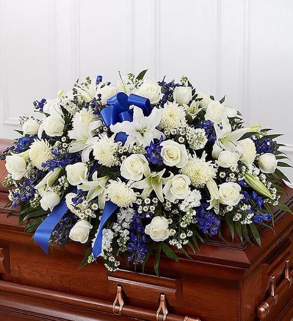Large white and blue floral casket spray with roses, lilies, and ribbon on a wood casket