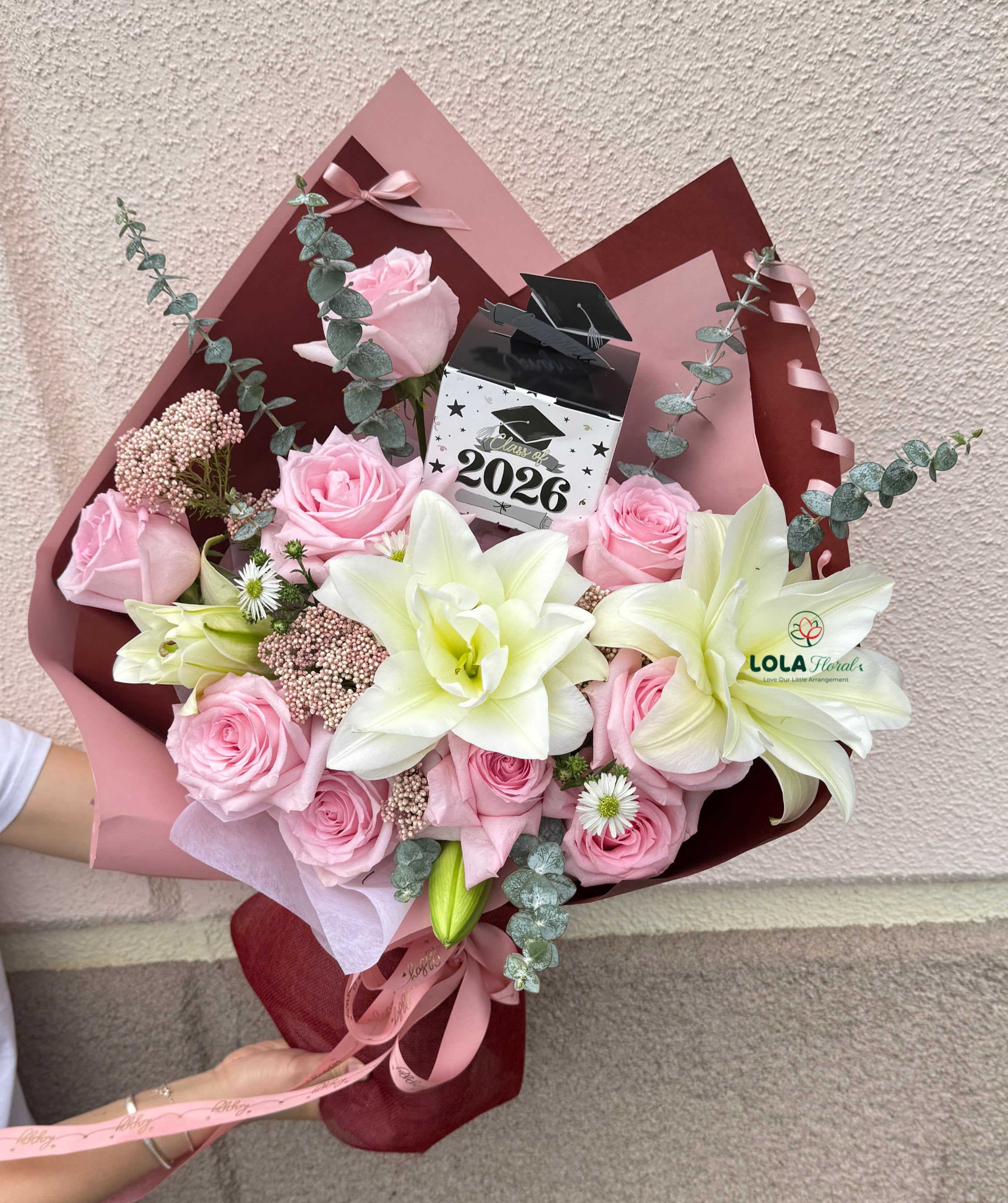 Graduation Surprise - Soft pink roses paired with large white lilies create a joyful, celebratory graduation bouquet designed to make the graduate feel truly celebrated. This graduation flower arrangement works beautifully as a congratulatory gift for commencement, a college send-off, or a proud high-school milestone-she'll receive a bouquet that reads like a personal standing ovation, complete with a "Class of 2026" topper.  The hand-tied arrangement is full and rounded, blending soft pink roses, showy white lilies, and delicate filler blooms in shades of pink and purple, all set against cool gray-green eucalyptus-like foliage for fresh contrast. It's wrapped in layered pink and burgundy decorative paper with satin ribbon accents and arrives in a gift box, so the presentation is polished and ready to give.  Perfect for the grad who loves pretty pastels or for family celebrating a major achievement, this bouquet sends a message of pride and joy. Lightly mist stems daily and recut if placing in water for longer enjoyment; choose delivery date at checkout to ensure it arrives for the ceremony or celebration.
