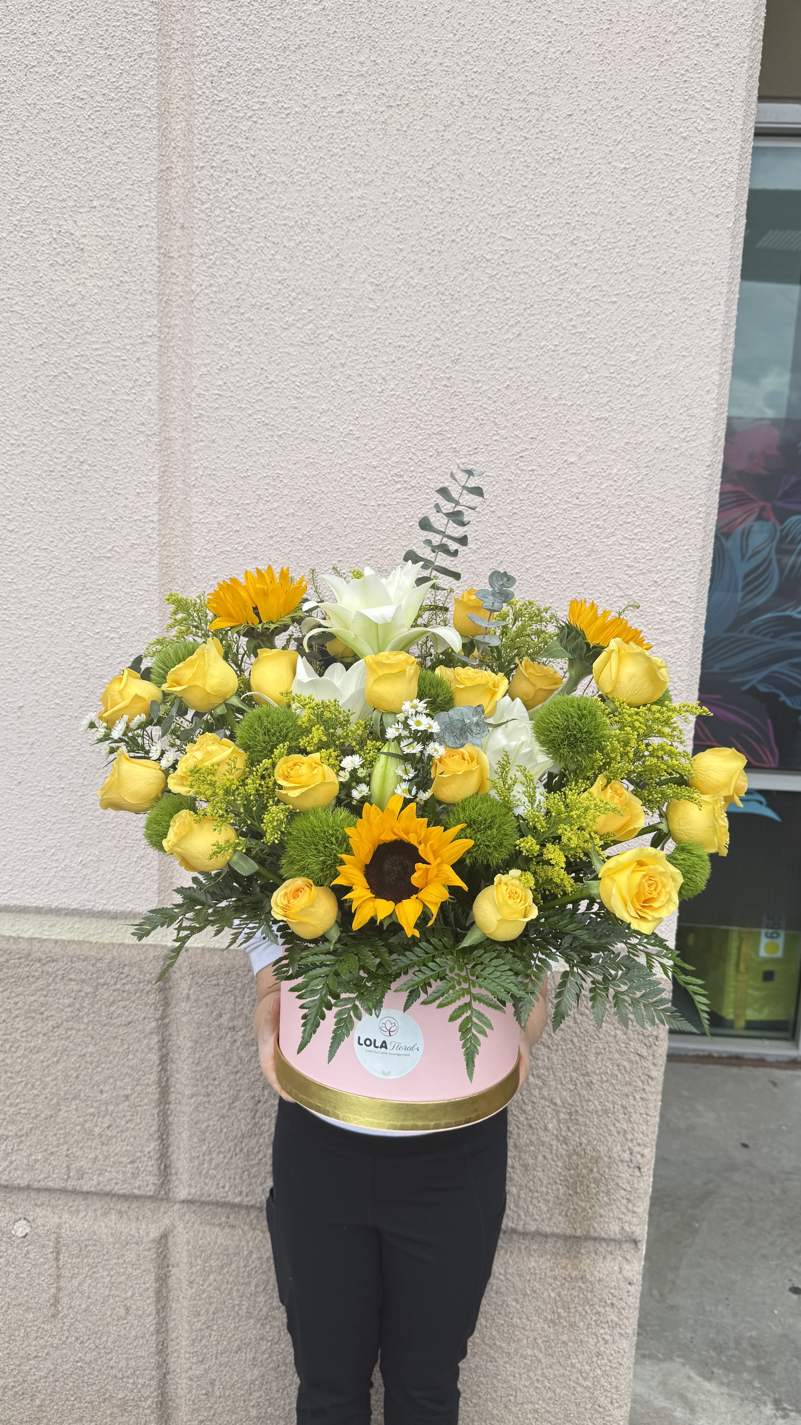 Hand wrapped box bouquet 01 - Sun-kissed and joyful, this flower arrangement in a pink round box is a cheerful pick-me-up for birthdays, congratulations, or a thoughtful "thinking of you" surprise. The rounded, dense composition places yellow roses and bold sunflowers front and center, with white lilies adding elegant height and a fresh, luminous contrast that will make the recipient feel celebrated.  A lively mix of small white daisy-like blooms and green textural accents fills out the design for a full, polished look. Presented in a pink cylindrical box with gold trim, the arrangement reads as an everyday gift with a touch of luxury-the sunny yellow, white, green, and pop-of-orange palette brightens any room and suits recipients who appreciate classic blooms with a playful twist.