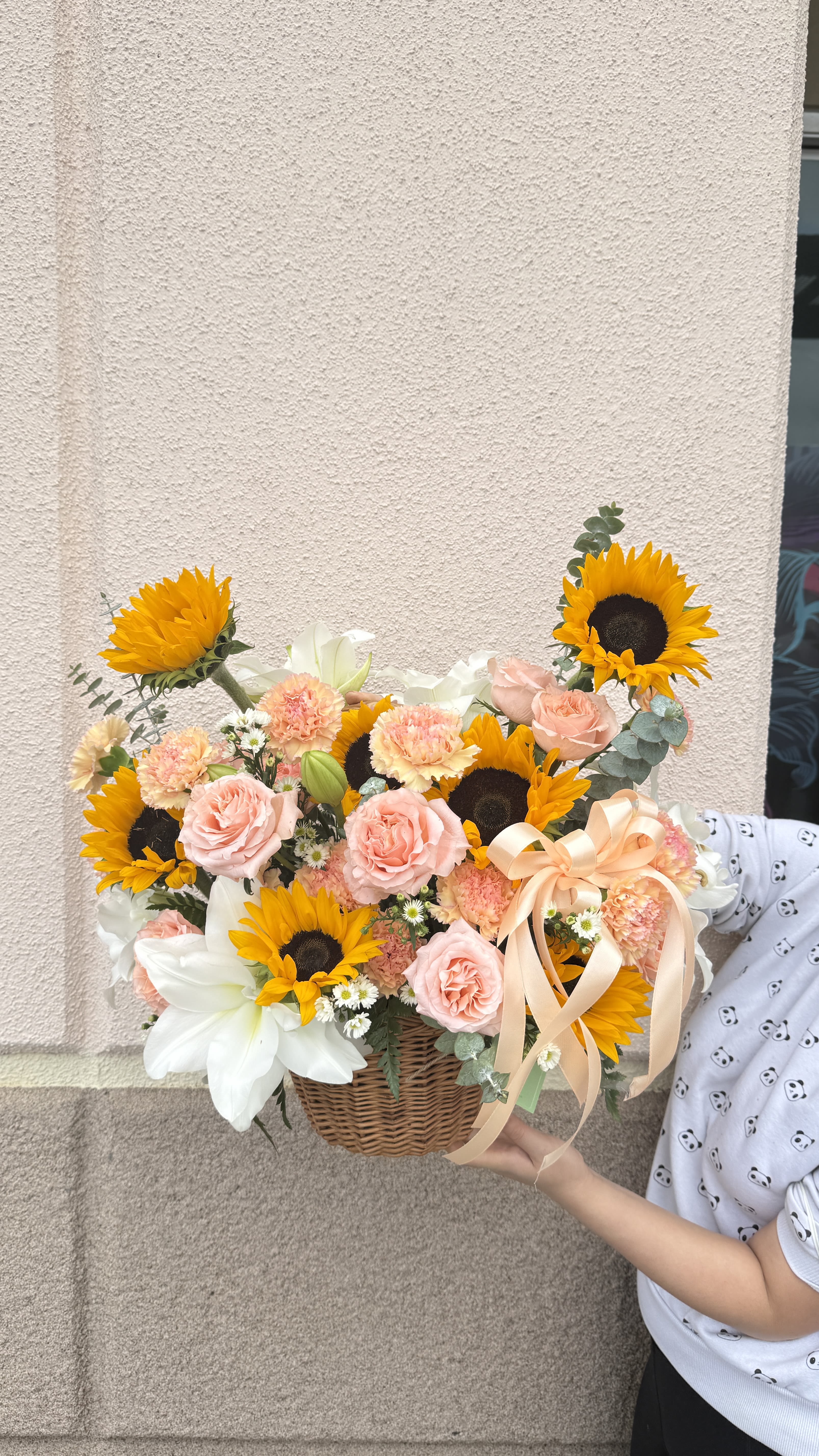 Hand wrapped box bouquet 02 - Bright sunflowers take center stage in this hand wrapped box bouquet 02, making it an uplifting birthday bouquet or just-because gift that sparkles with sunny cheer. The arrangement's mix of pink roses, white lilies, peach carnations and cheerful daisies creates a warm, celebratory feel your person will notice the moment it arrives - the kind of bouquet that makes someone feel truly seen and appreciated.  Packed tightly in a woven wicker basket and finished with a large peach ribbon, the composition reads full and rounded with a pleasing contrast between vivid yellow centers and soft pastel blooms. Expect a generous, garden-fresh look with textural variety from smooth rose petals, ruffled carnations, and sculpted lily blooms. Ideal for birthdays, thank-you gestures, or brightening an everyday moment; standard care is to keep flowers in fresh water and trim stems every few days to extend their life.