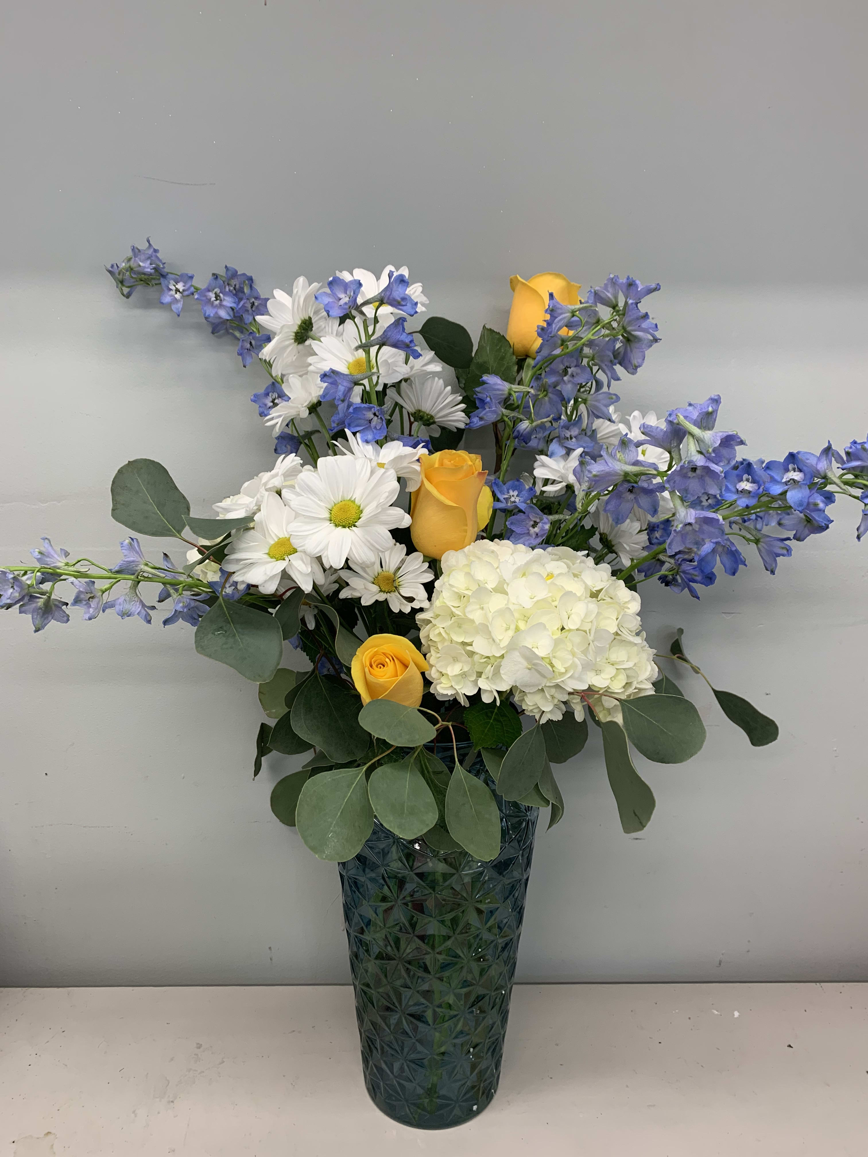 Yellow roses, white daisies, and blue flowers in a tall glass vase