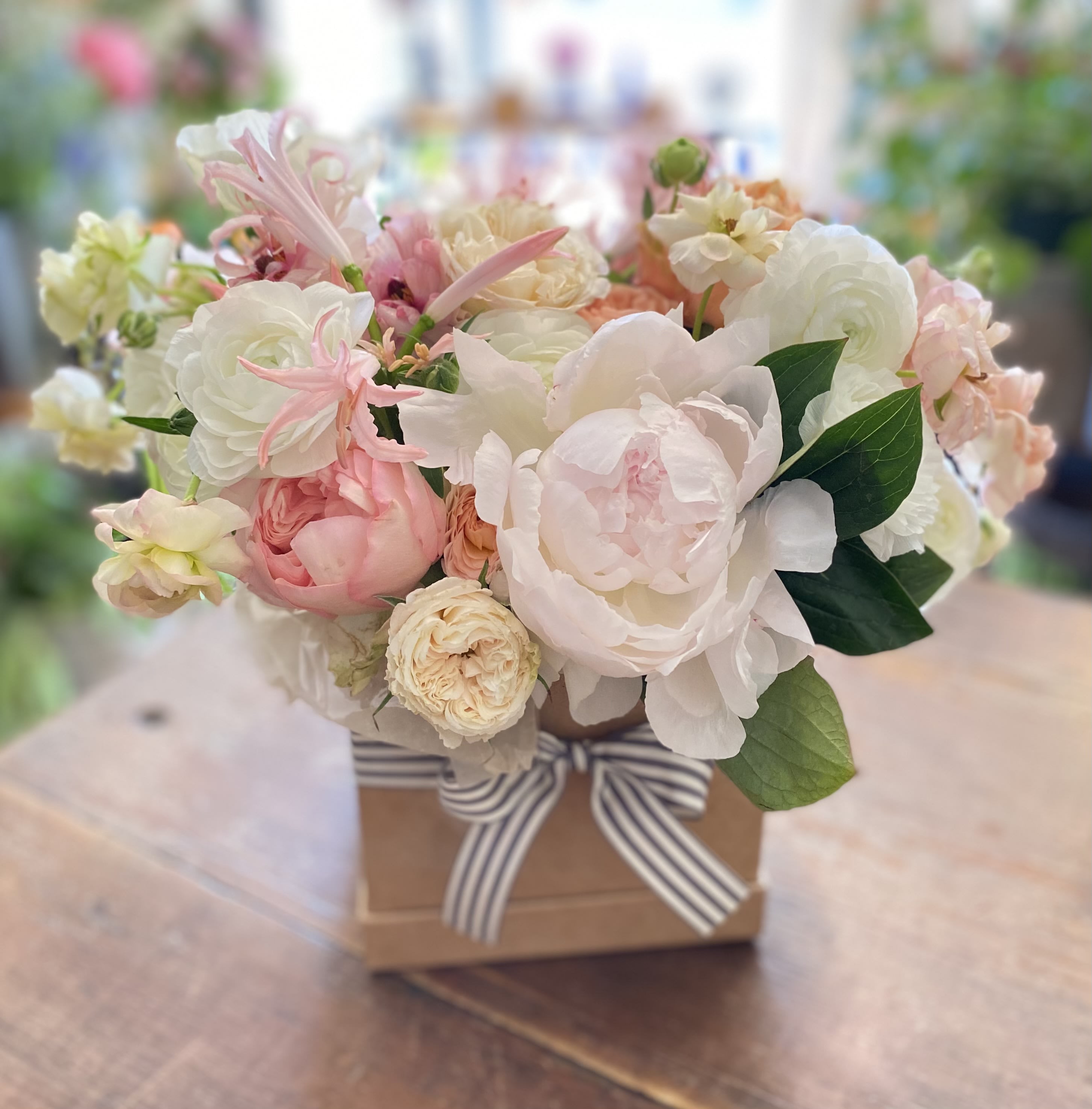Pastel pink and white peonies and roses arranged in a kraft box with striped ribbon bow