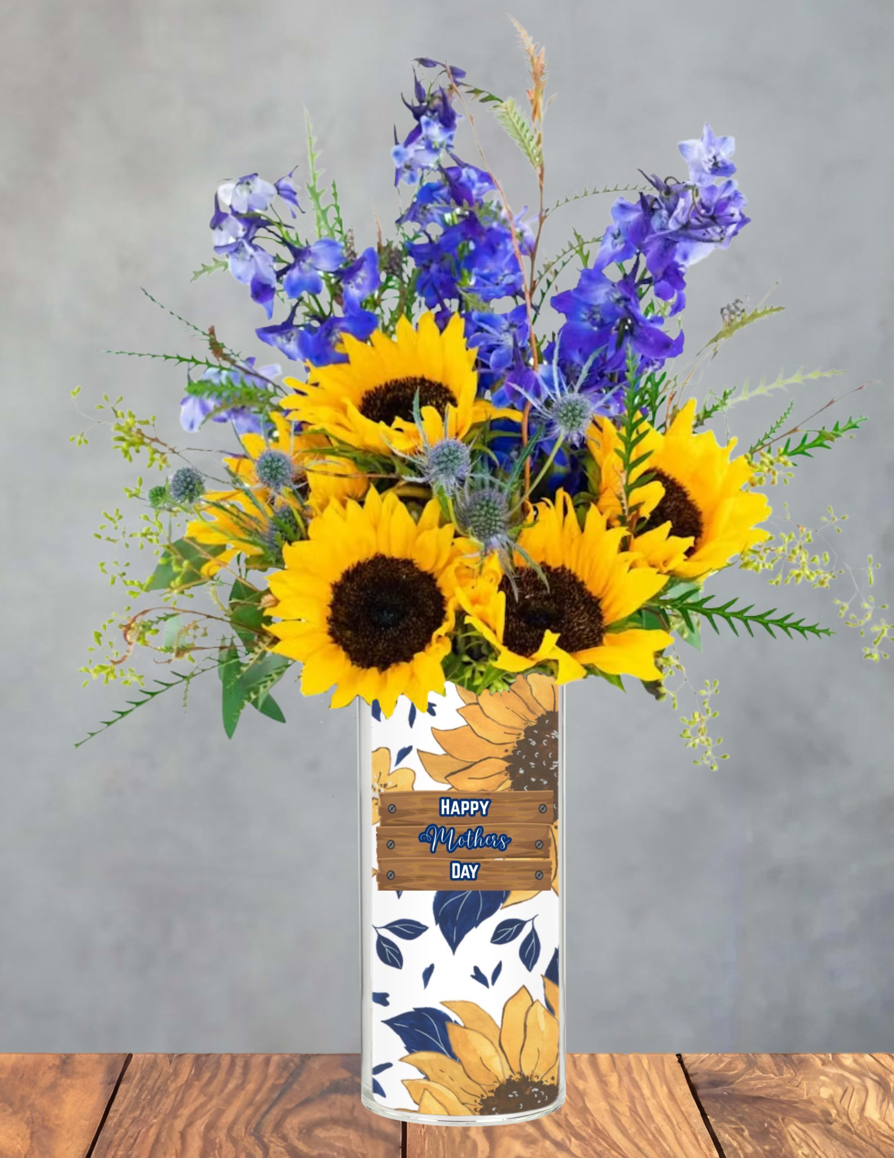Moms sunny skies - Bright yellow sunflowers paired with tall blue delphinium-like spikes make a joyful Mother's Day bouquet that instantly brightens a room - perfect as a Mother's Day gift or a cheerful thank-you arrangement. The upright, abundant silhouette and crisp spring palette of yellow, blue, and white create an energetic contrast that will make the person you're thinking of feel celebrated and seen.  This arrangement is presented in a tall clear glass vase wrapped with a decorative Mother's Day floral label and finished with a small wooden-style "Happy Mothers Day" tag. The sunflowers provide bold focal blooms while the delphinium-like stems add height and airy texture; touches of white and other filler flowers soften the composition for a balanced, fresh look. The tall vase keeps stems elegantly supported and displays the arrangement beautifully on a table or mantle.  Ideal for moms, grandmothers, or anyone who loves sunny, uplifting flowers, this bouquet conveys gratitude and warmth without being overly formal. Care is simple: keep the water fresh, recut stems every few days, and place away from direct heat to prolong bloom life - perfect for gifting and enjoying all week long.
