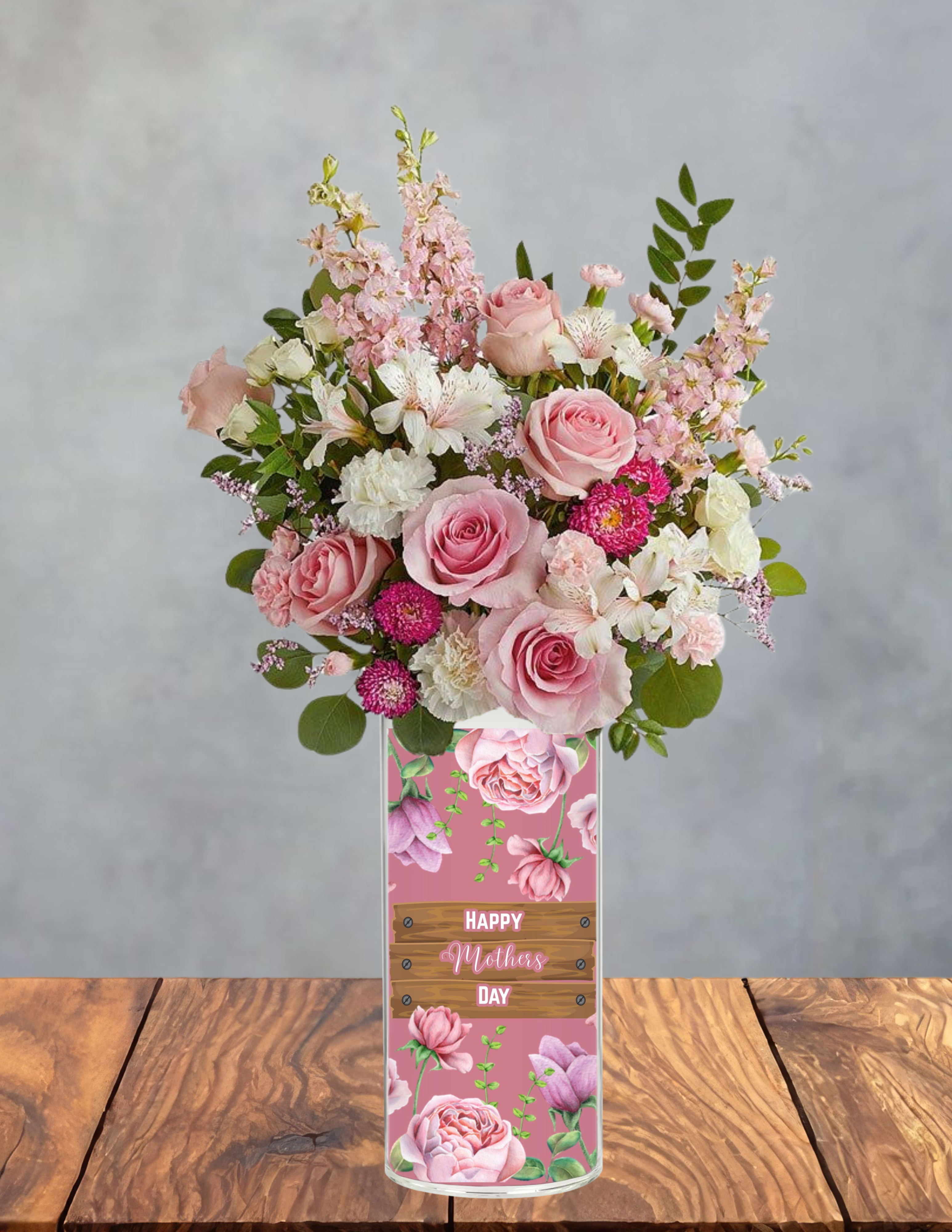 Mamas pink delight - Soft pink roses and airy pink stock come together in a tender Mother's Day bouquet designed to make Mom feel truly cherished. This flower arrangement for Mother's Day mixes roses, white alstroemeria, soft carnations, petite chrysanthemums, and touches of purple-magenta accents to create a sentimental, cheerful presentation that reads feminine and heartfelt.  Arranged densely in a tall clear glass vase, the flowers form a lush, rounded silhouette with delicate texture from the stock and small chrysanthemum blooms. The vase features a decorative wood-plank style Mother's Day label and a pink graphic, making it ready to set on a kitchen table or bedside dresser. Colors include pale pinks and whites grounded by green foliage and bright magenta pops for contrast.  Perfect for celebrating Mom, a new mother, or any woman who loves classic, romantic florals, this bouquet says "you're appreciated" in soft, floral tones. Vase dimensions and care: keep water fresh, recut stems every few days, and place out of direct heat to extend bloom life; the arrangement arrives gift-ready with the Mother's Day label already attached.