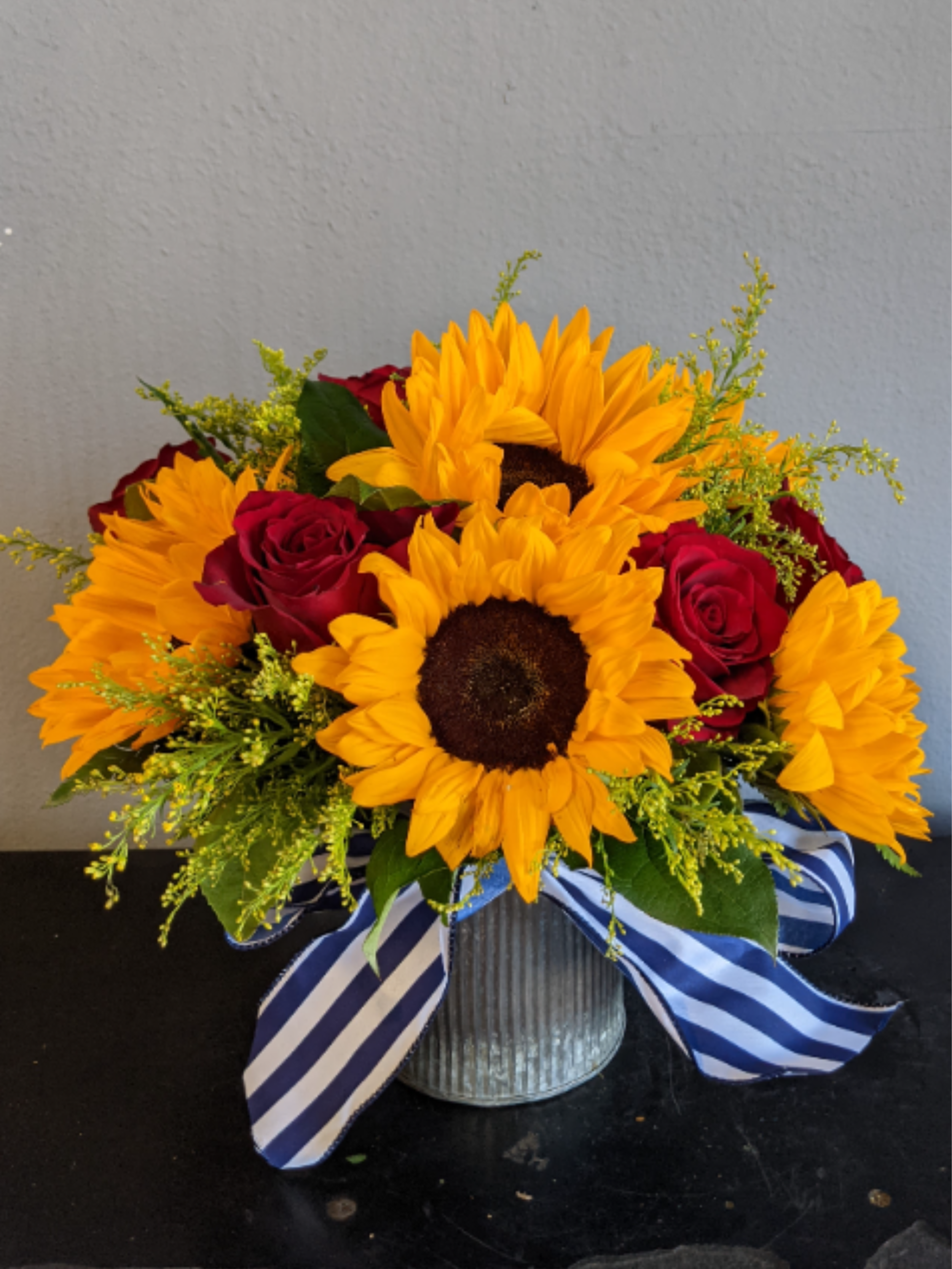 Low arrangement of bright sunflowers and red roses in a metal vase with a striped blue ribbon.