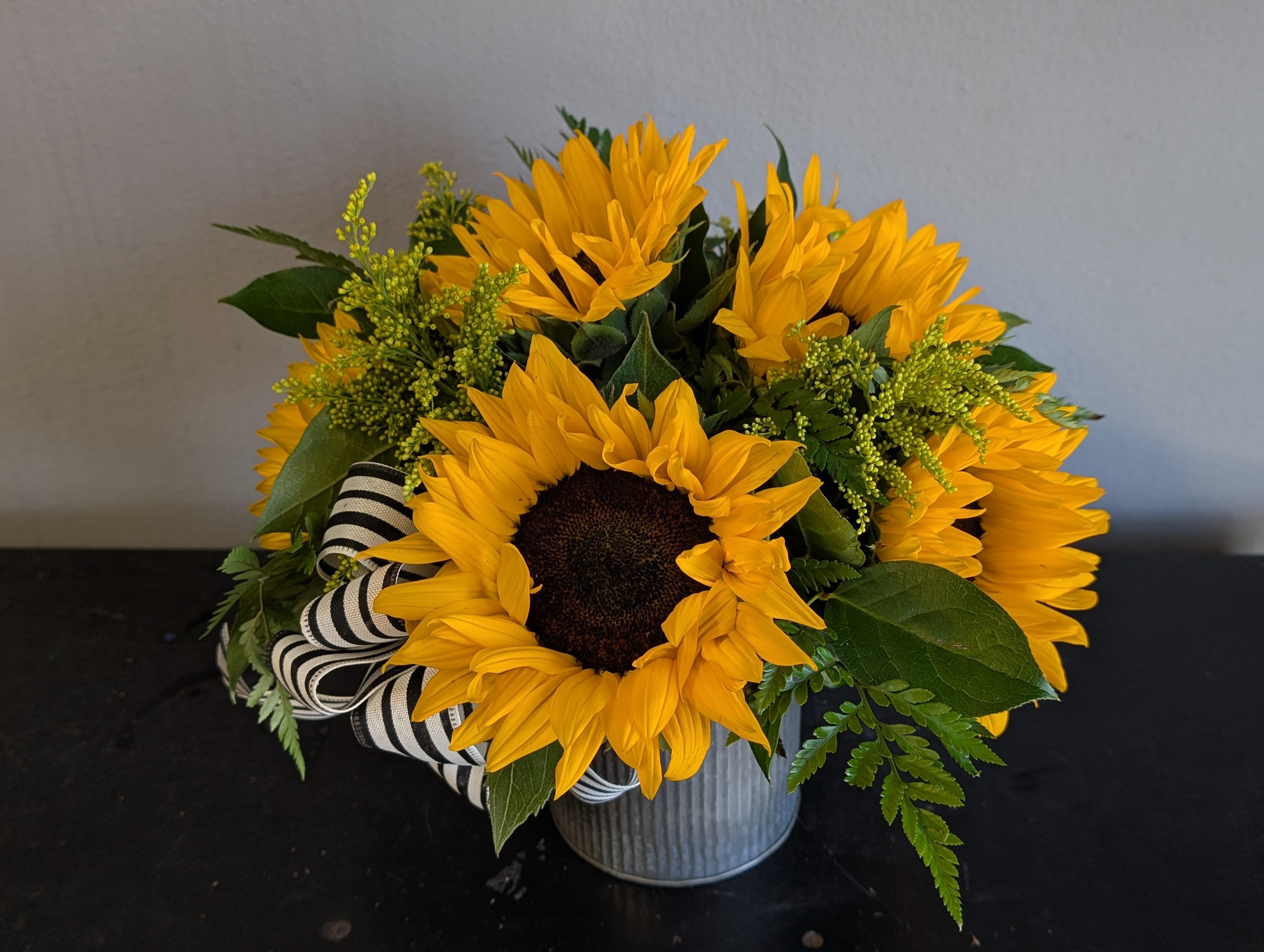 Short arrangement of yellow sunflowers in a metal pot with a black and white striped ribbon.