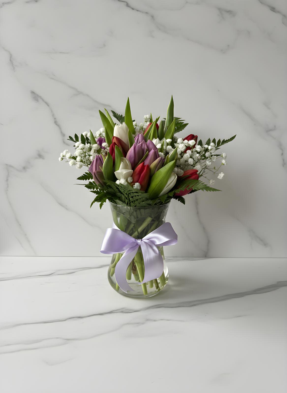 20 Tulips Vase - 3 Colors Designer's Mix Choice  - Celebrate the beauty of the season with our Spring Garden Tulip Arrangement, a fresh and elegant design featuring a charming mix of colorful tulips, delicate baby’s breath, and lush greenery. Arranged in a classic clear glass vase and finished with a soft satin ribbon, this bouquet brings a natural garden-inspired look to any space.  The soft combination of pastel and vibrant tulip tones creates a bright and uplifting arrangement perfect for birthdays, thank-you gifts, spring celebrations, or simply brightening someone’s day.  Each arrangement is carefully handcrafted to ensure a balanced, fresh, and elegant presentation.  Please note: Tulip colors and flower varieties may vary slightly depending on seasonal availability while maintaining the overall style and fullness of the arrangement.